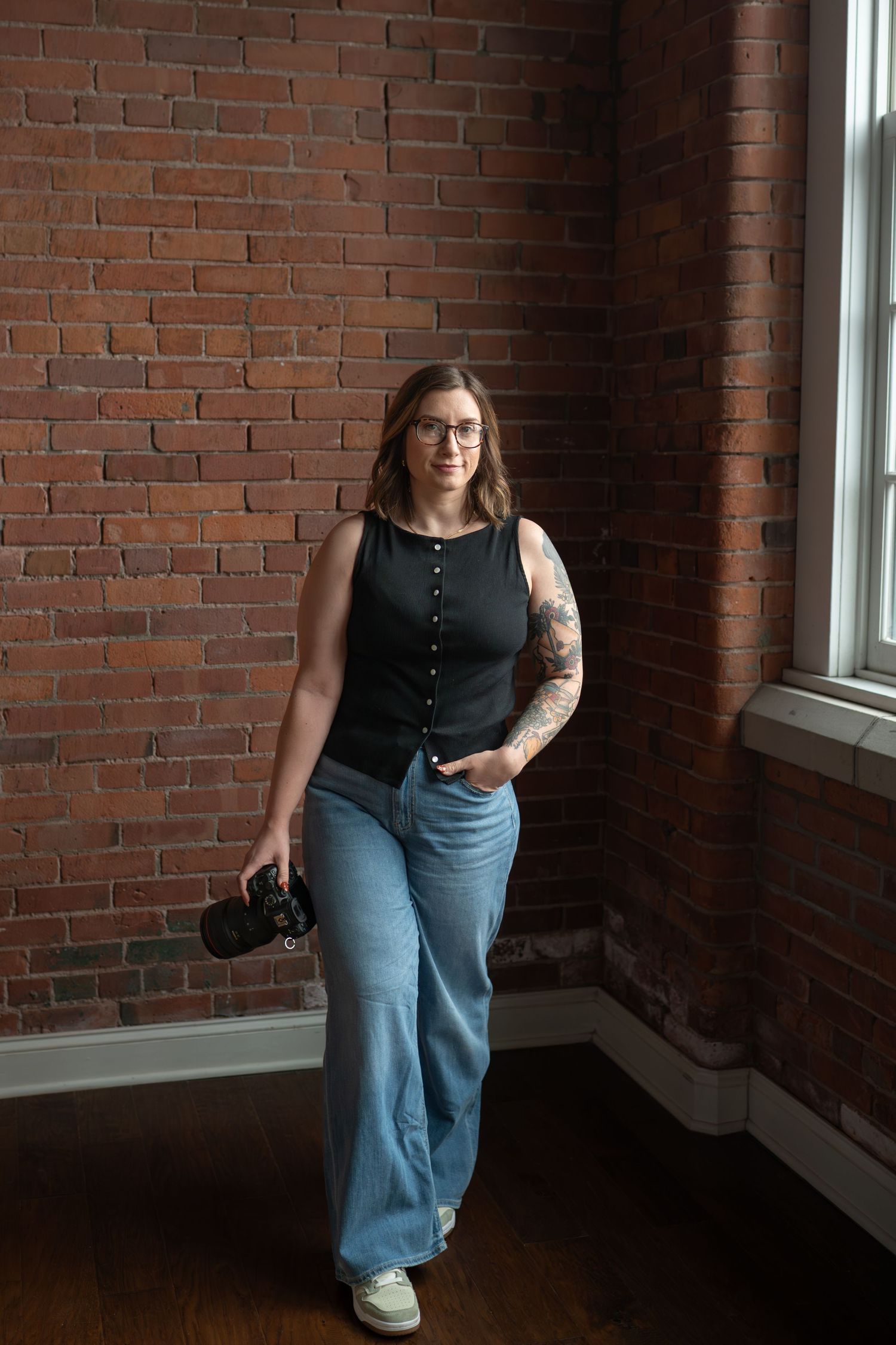 A tattooed woman in glasses holds a camera, wearing a black tank and wide-leg jeans, standing by a brick wall.