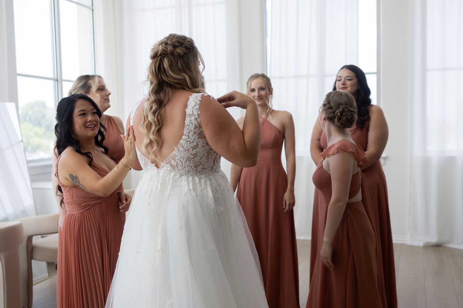 Bride in lace wedding gown surrounded by bridesmaids in dusty rose dresses helping her get ready in bright room.