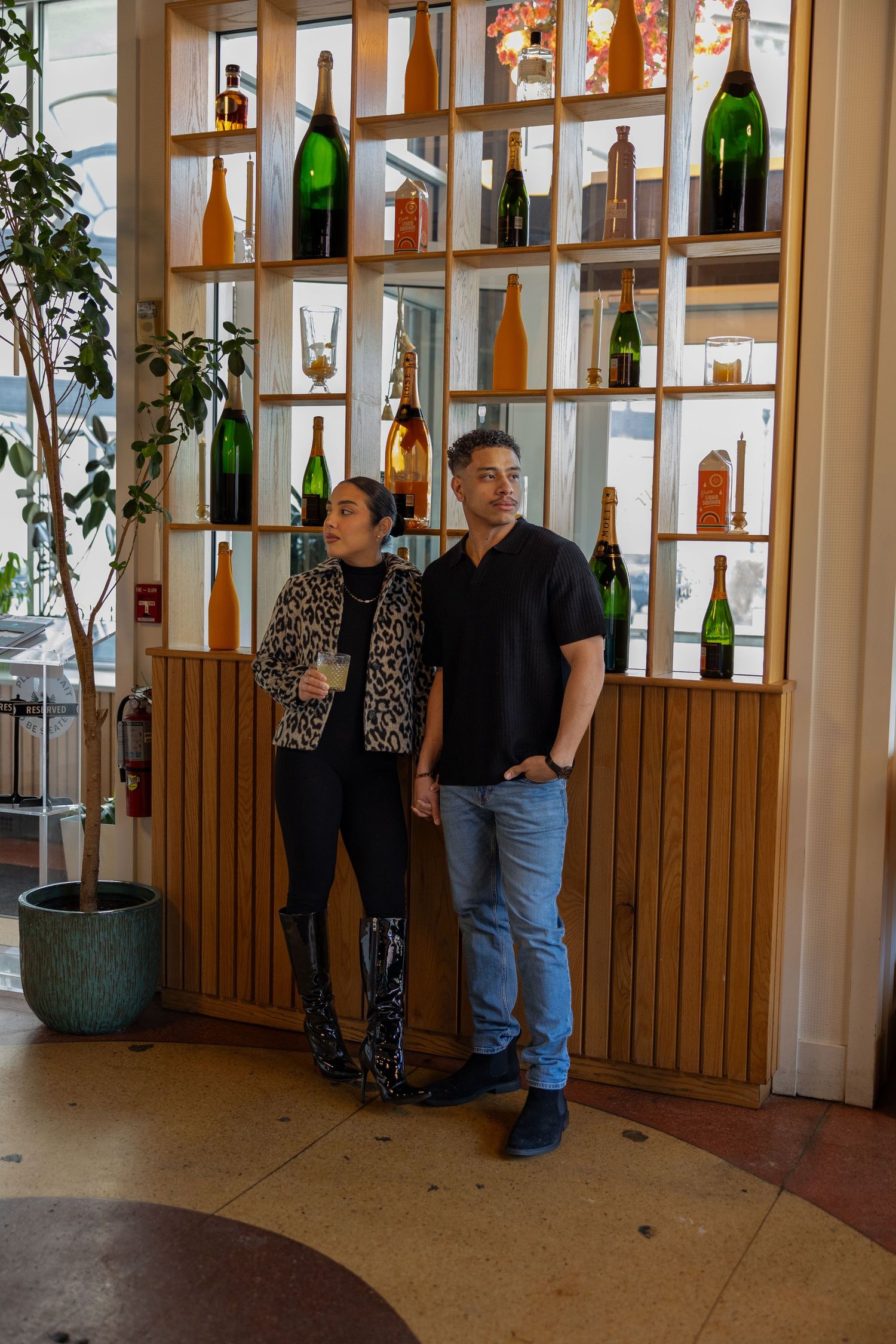 Couple posing in front of a wooden shelving display with large wine bottles in an upscale restaurant.