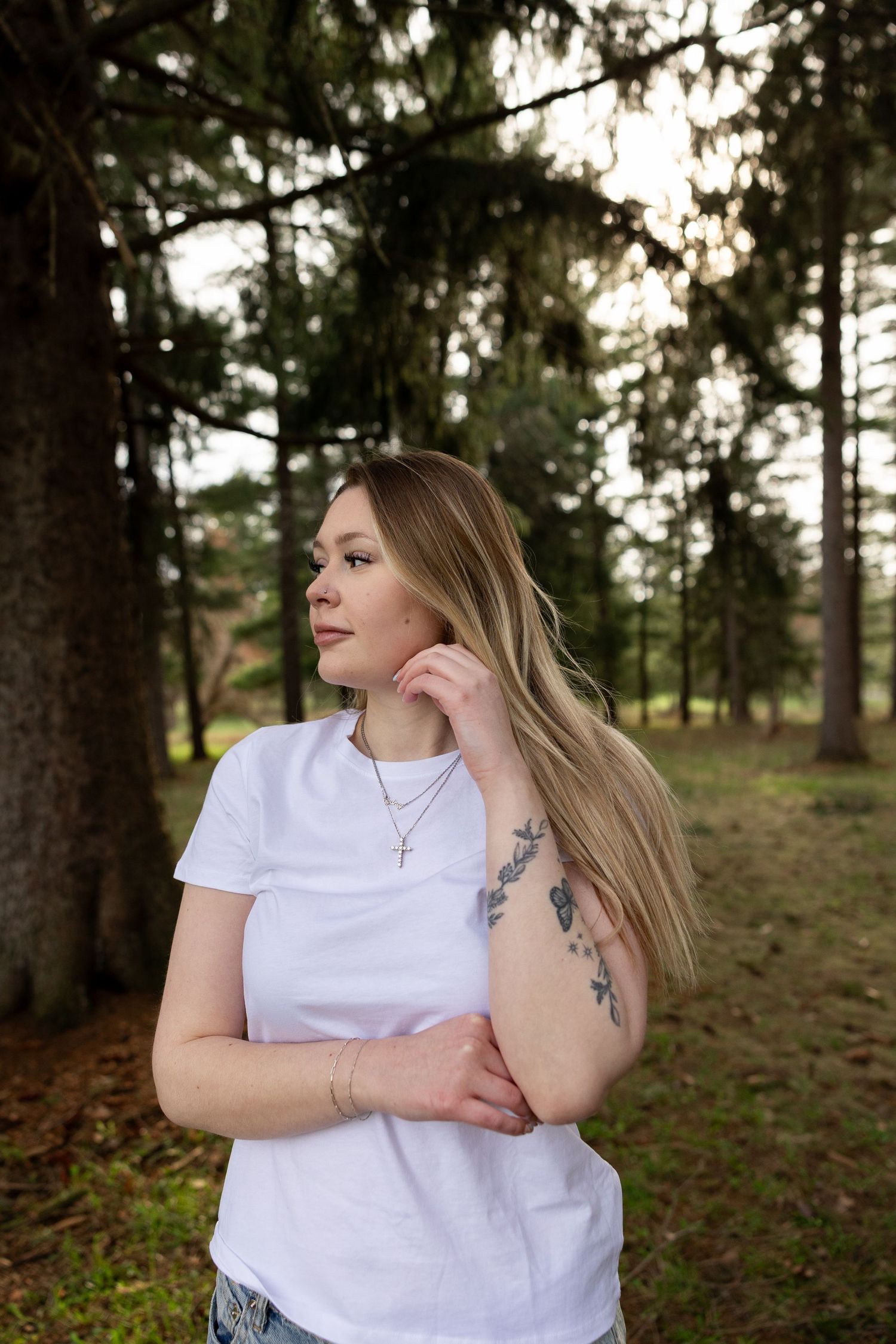 Young blonde woman with tattoos in white t-shirt and cross necklace posing outdoors in a pine forest.