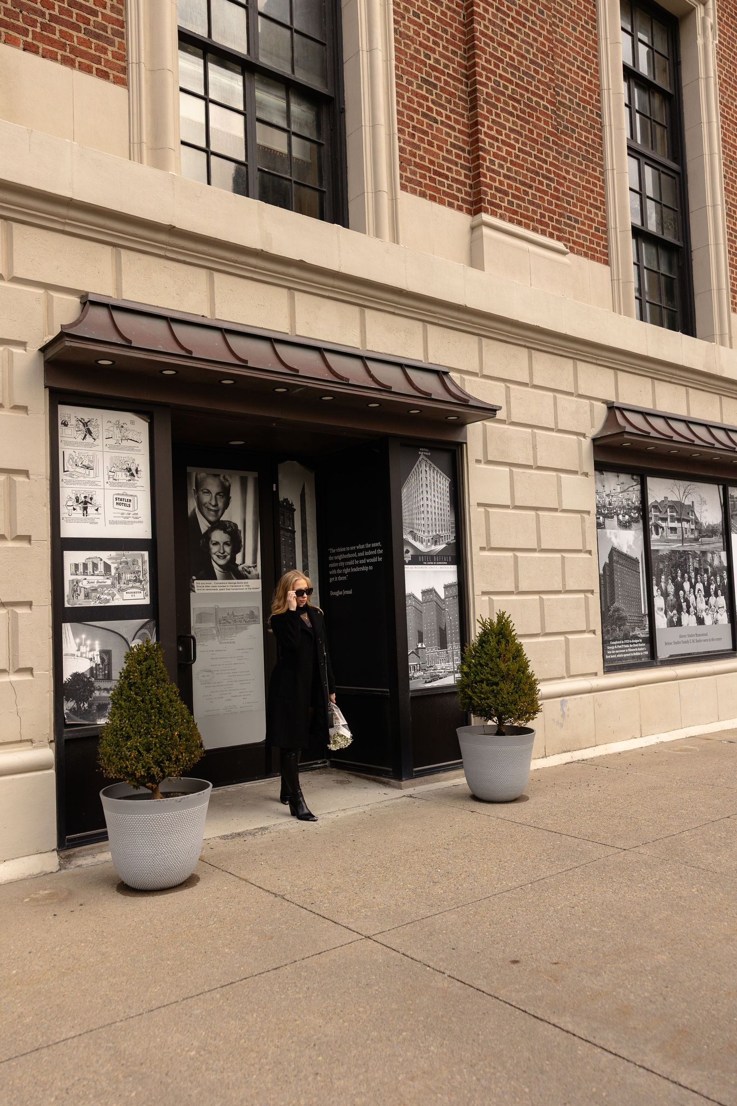 Woman in black outfit standing outside a brick museum building with historical photo displays and potted shrubs.