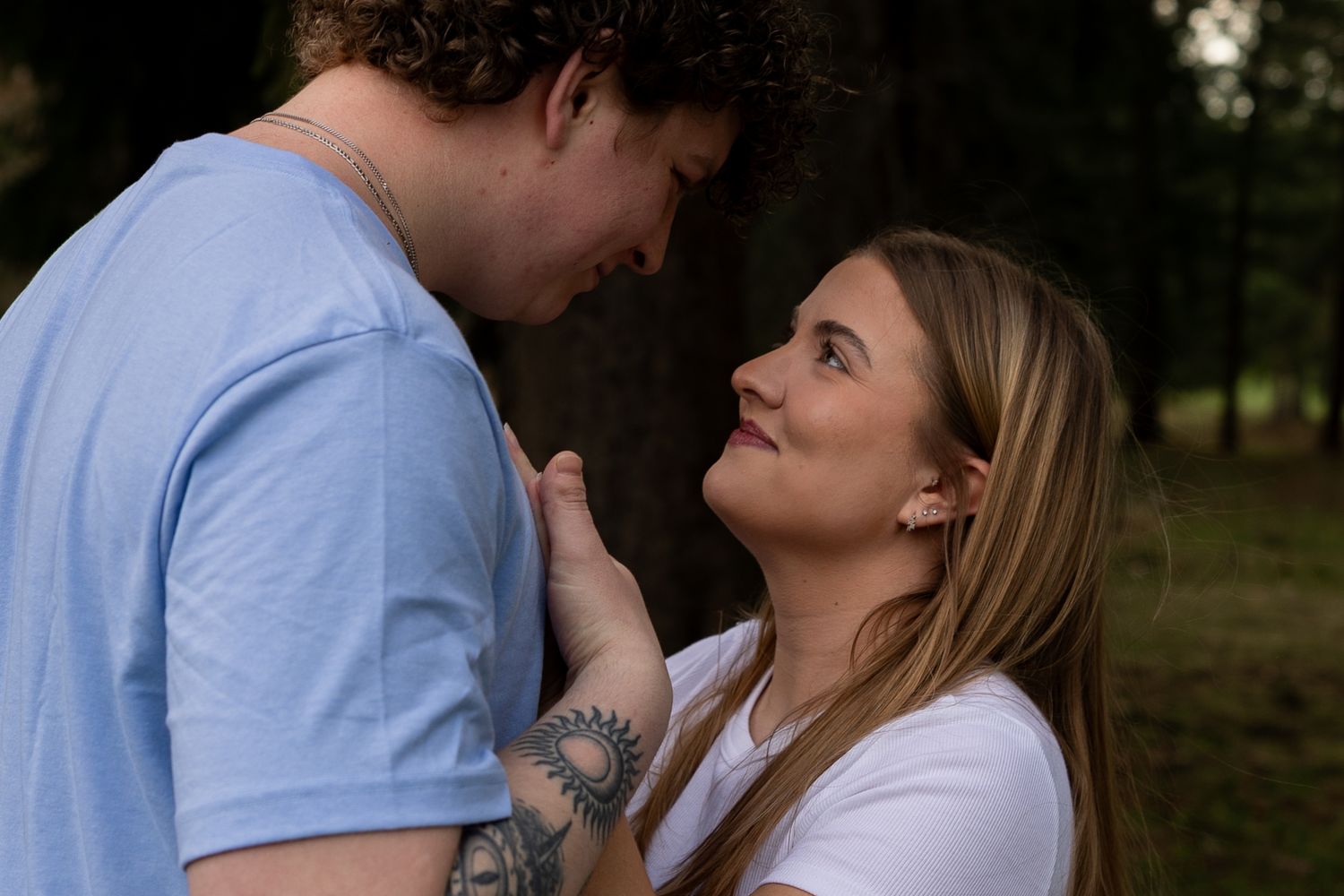 A couple shares a tender moment outdoors, woman smiling up at tattooed man in blue shirt among trees.