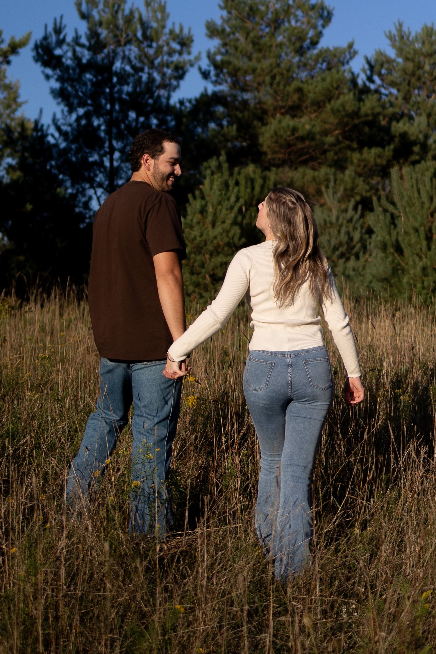 A couple holds hands while walking through a tall grass field with pine trees in the background on a sunny day.
