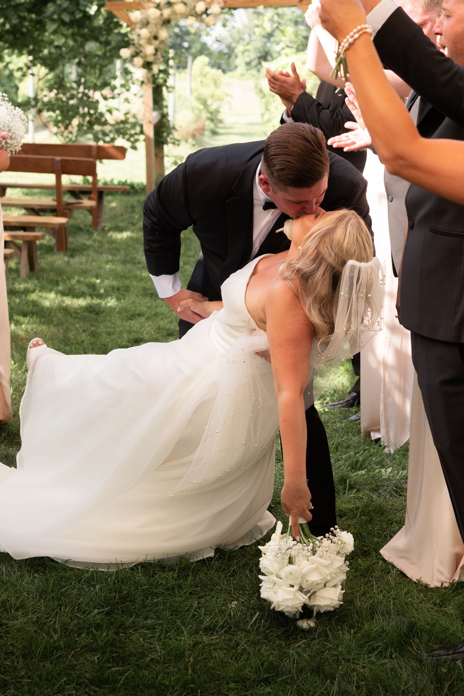 Bride and groom share a romantic dip kiss during outdoor wedding ceremony on green lawn, bride holding white rose bouquet.