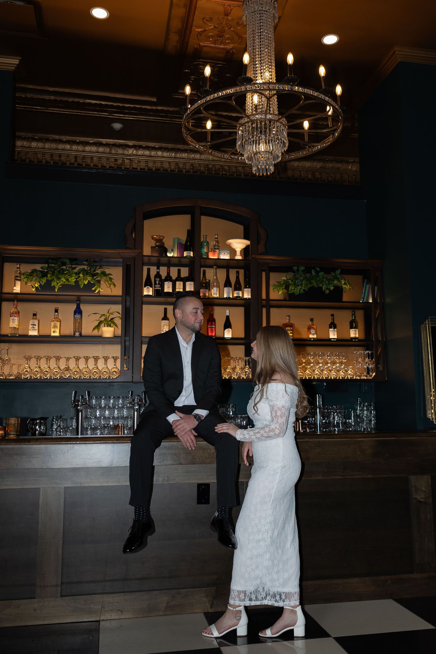Elegant couple in a dark upscale bar, man in suit sitting on counter, woman in white lace dress, chandelier above.