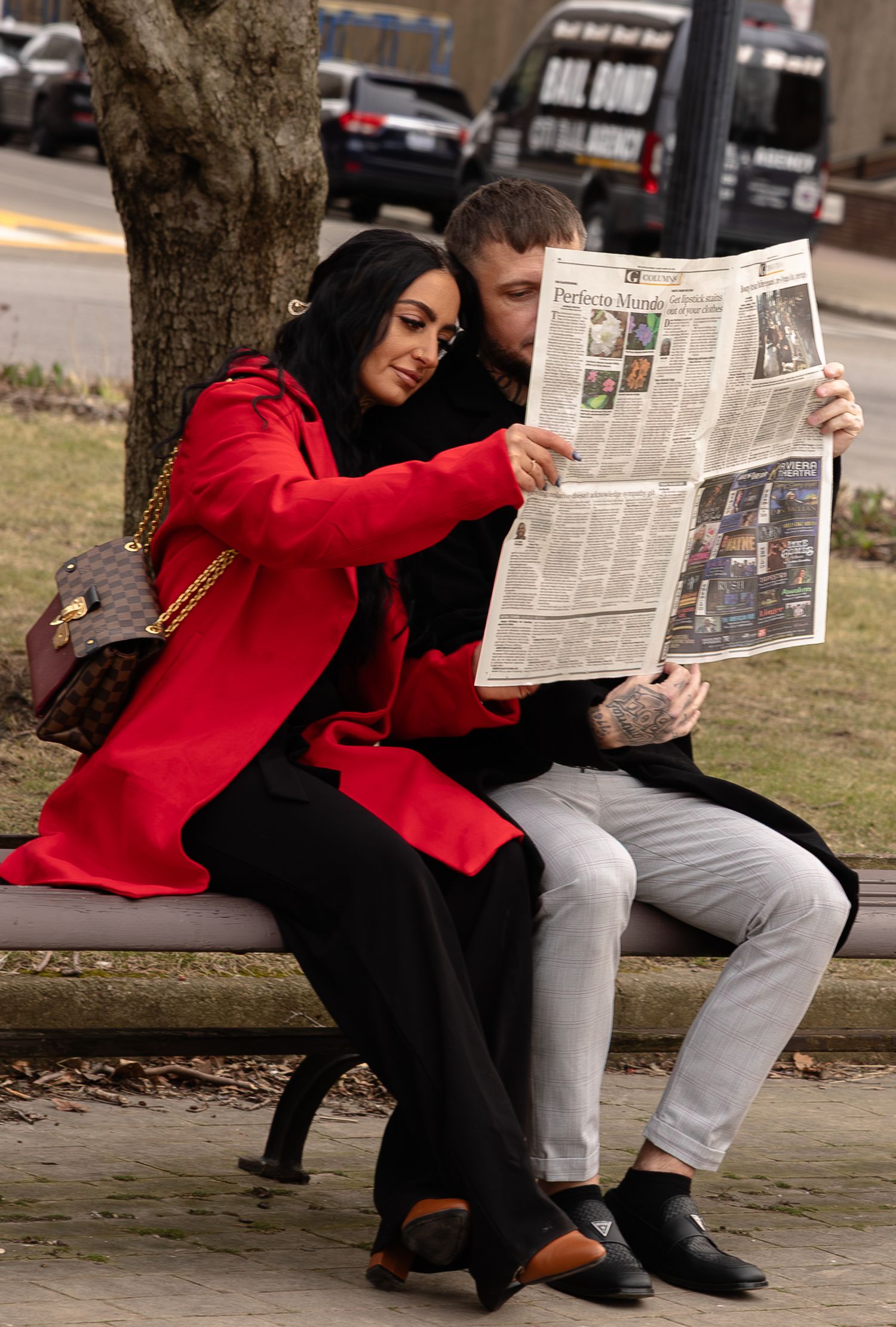 Couple sitting on a park bench reading a newspaper together, woman in red coat with Louis Vuitton bag.