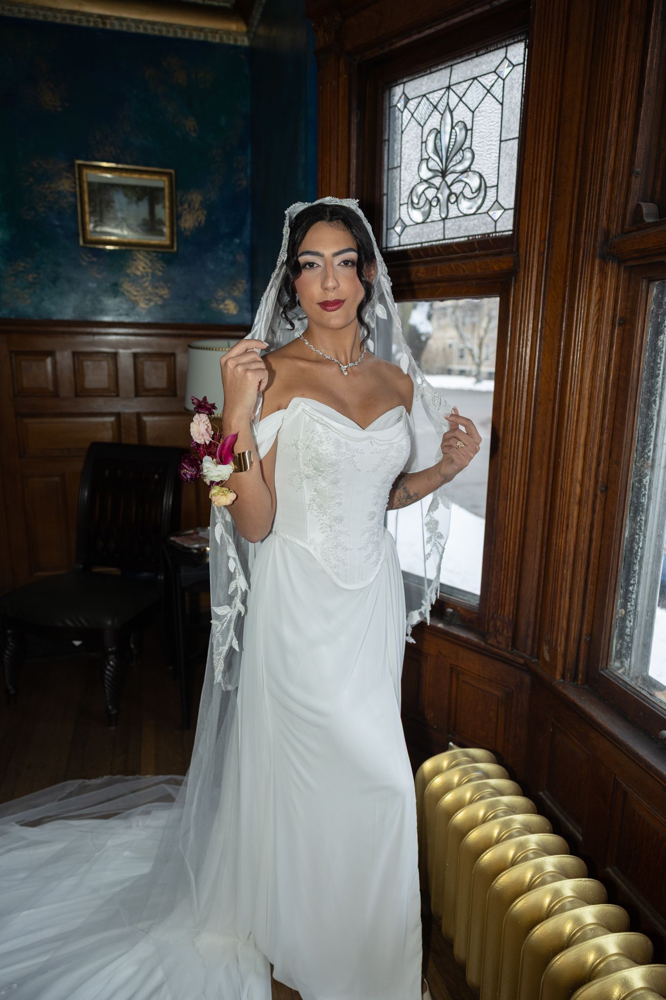 Elegant bride in white corset gown and veil holds bouquet by stained glass window in ornate wood-paneled room.
