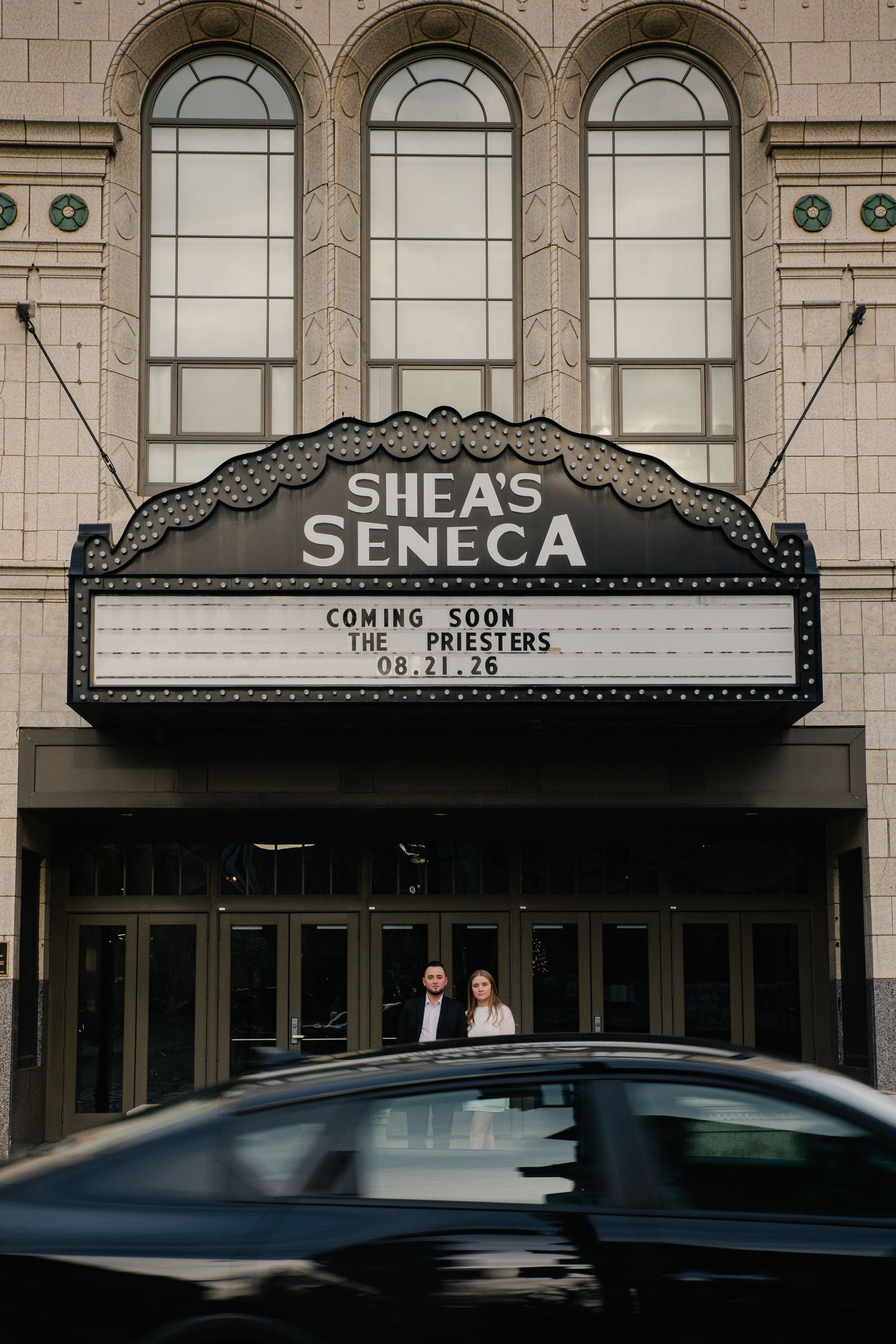 Couple poses under Shea's Seneca Theatre marquee reading The Priesters coming soon 08.21.26 in Buffalo NY.