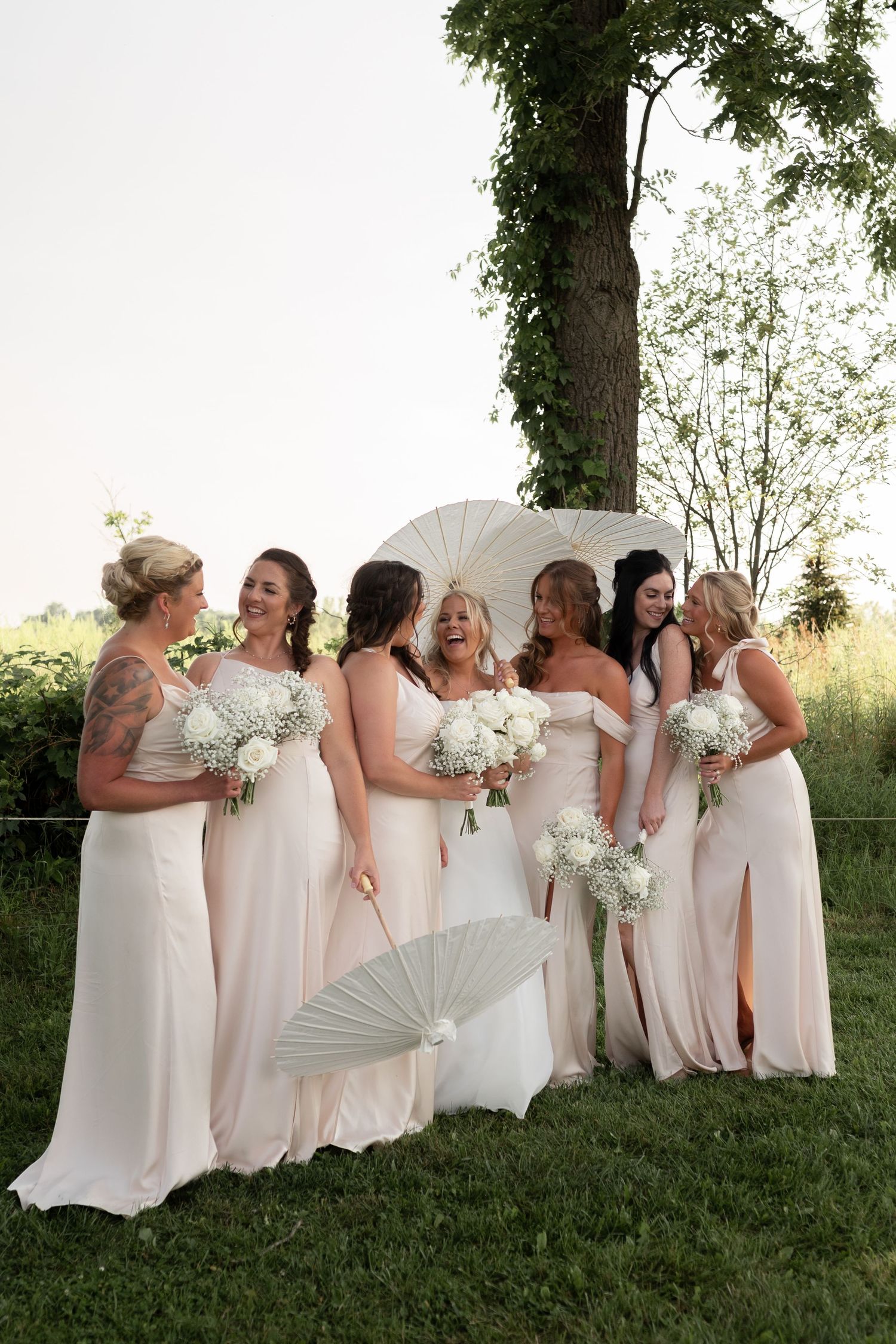 Bride and bridesmaids in blush gowns holding white bouquets and parasols laughing outdoors at a summer wedding.