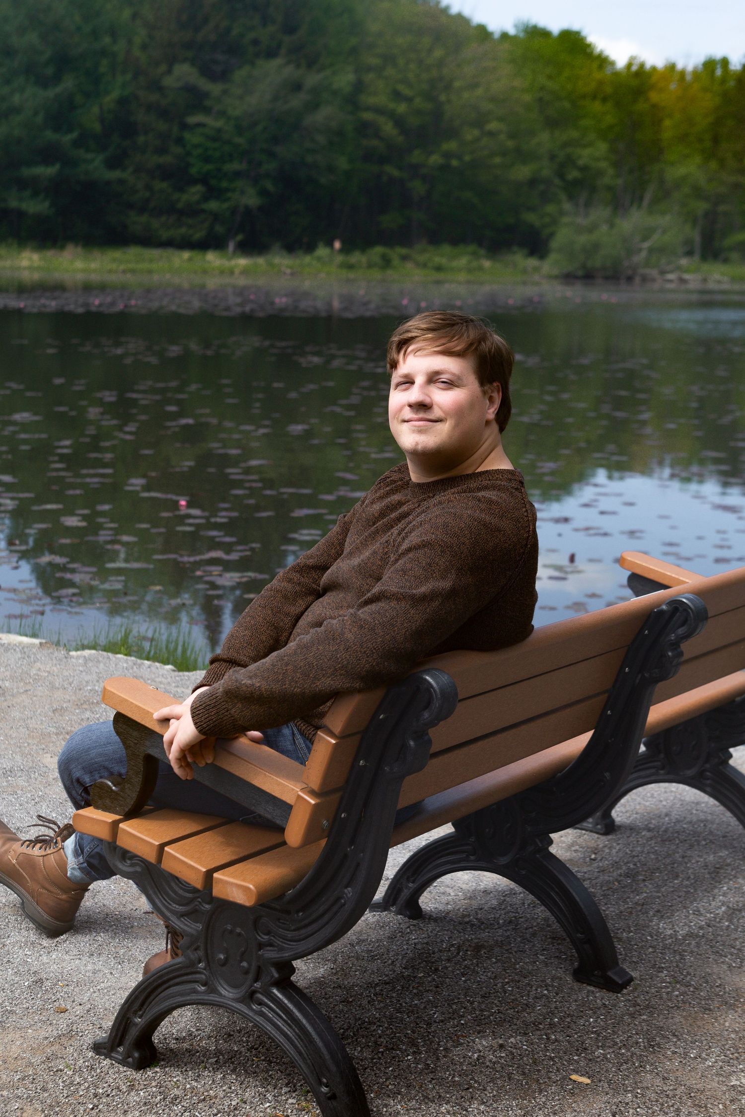 Young man in brown sweater sitting relaxed on a wooden park bench beside a calm reflective lake surrounded by green trees.