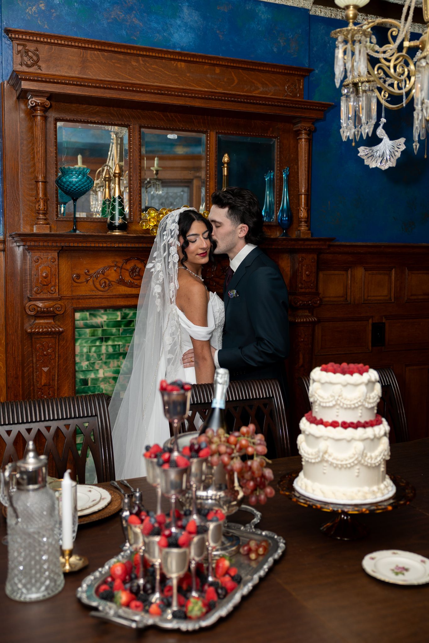 Bride and groom share a romantic moment beside a three-tier wedding cake with champagne and fruit on elegant table.