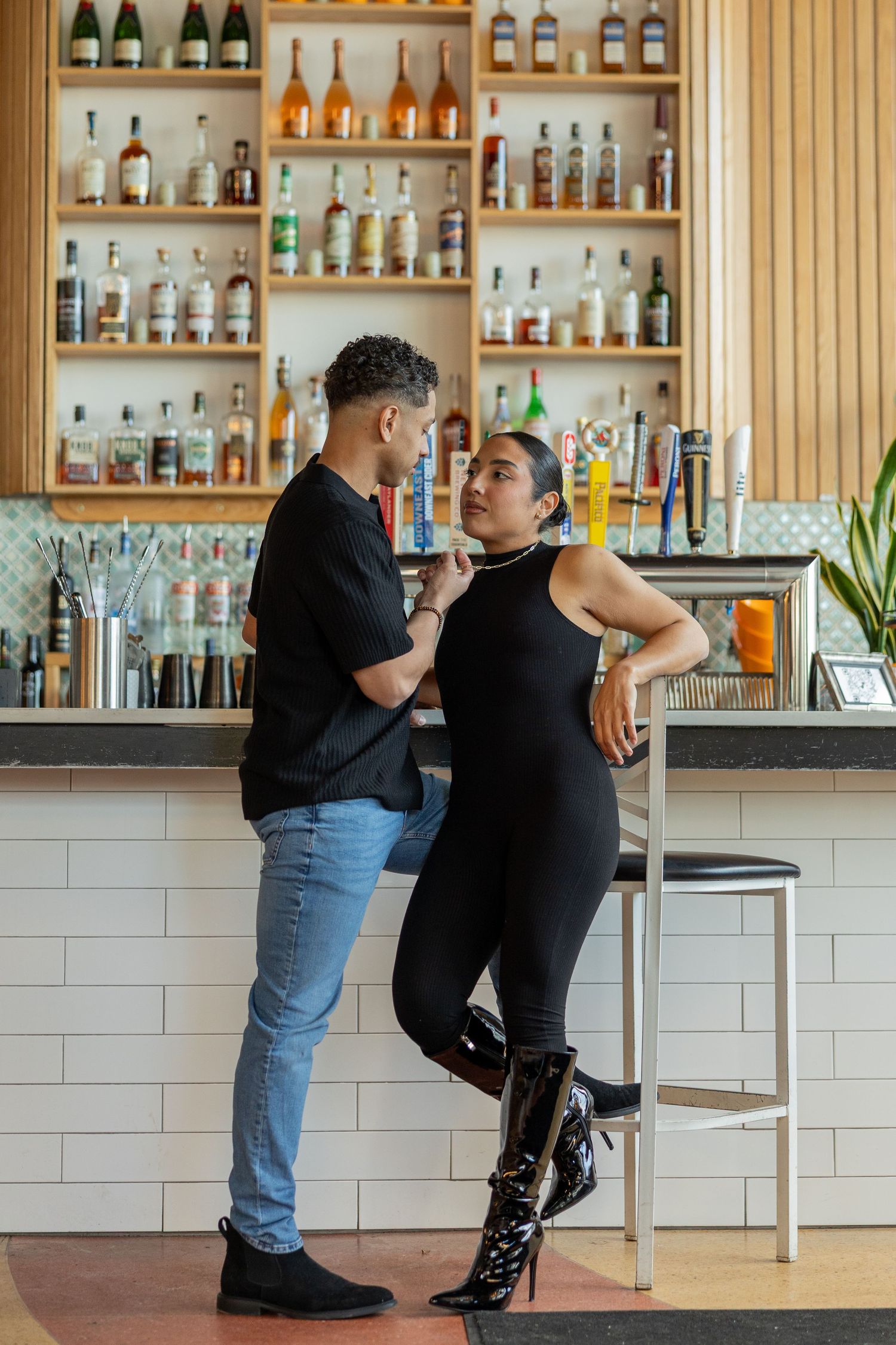Couple in black outfits sharing a romantic moment at a stylish bar with shelves of liquor bottles.