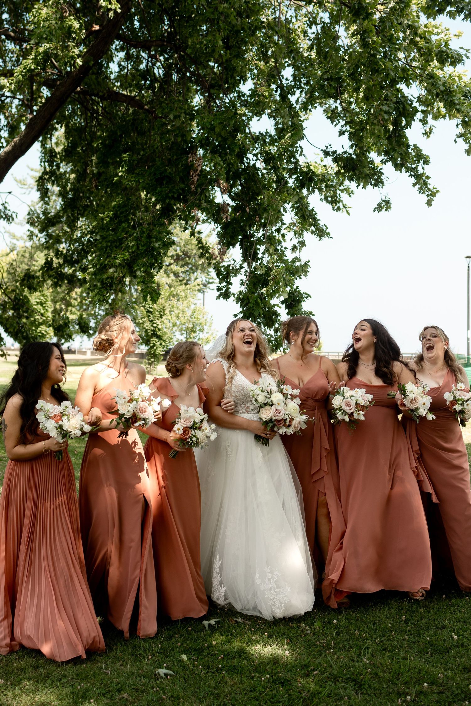 Bride in white gown laughing with bridesmaids in rust orange dresses holding bouquets outdoors under green tree.