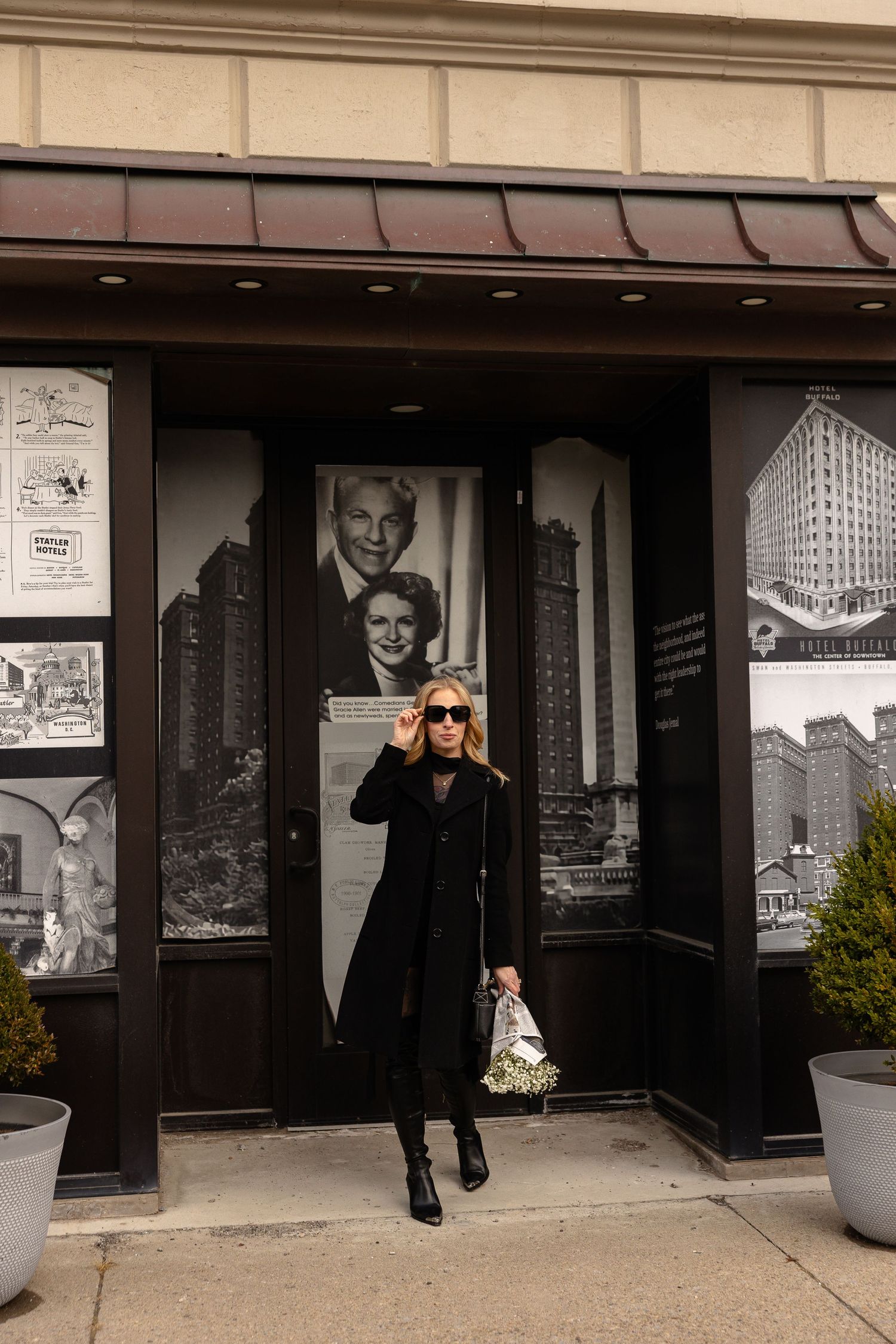 Stylish woman in black coat and sunglasses posing in front of vintage black and white photo display on building exterior.