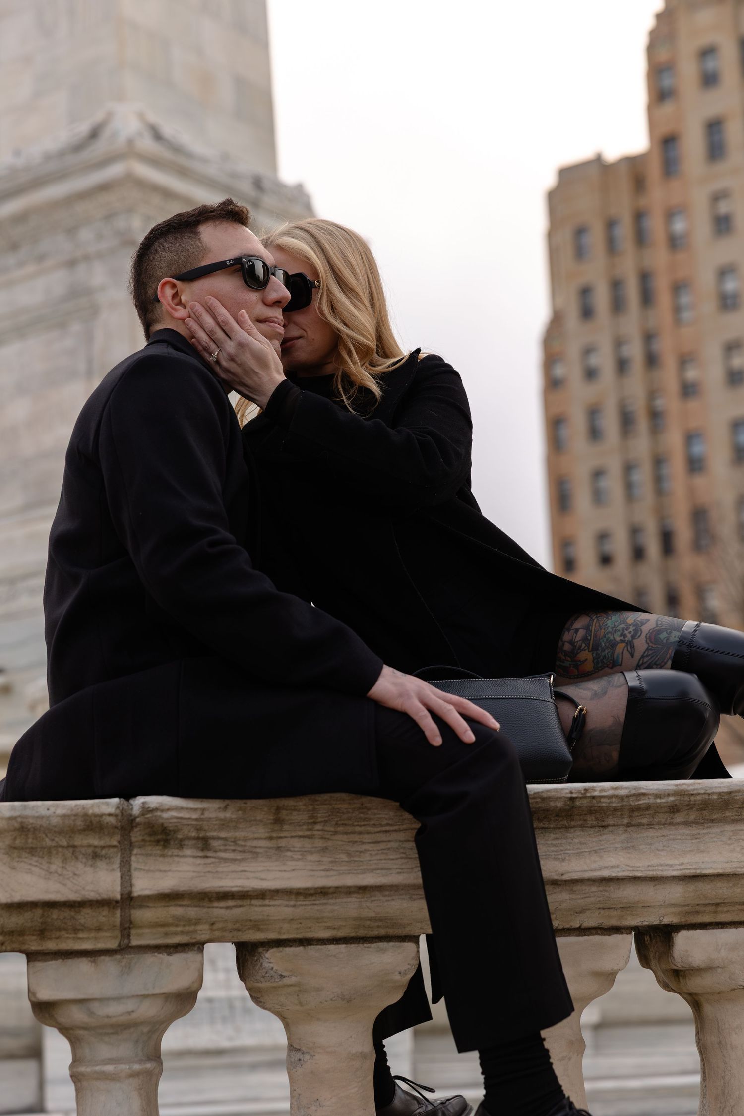 Couple in black outfits sharing a kiss while sitting on a stone balustrade with city buildings in the background.