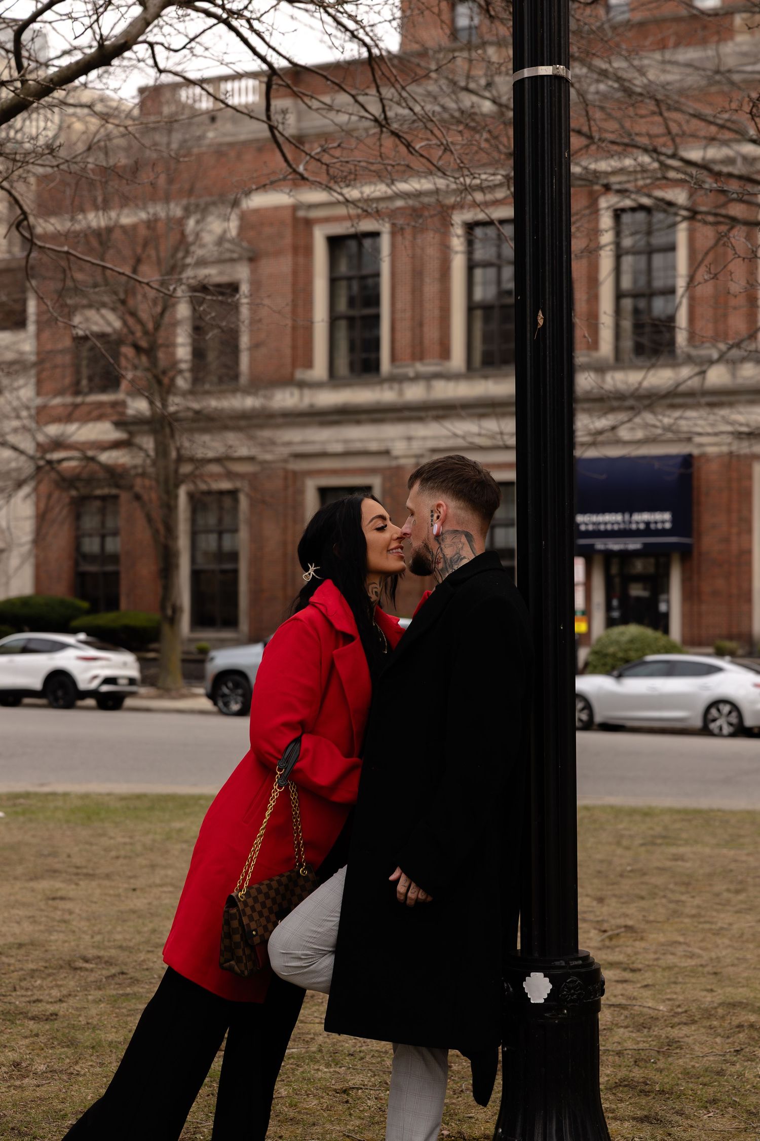 Couple kissing by a black street lamp in a city park, woman in red coat holding Louis Vuitton bag.
