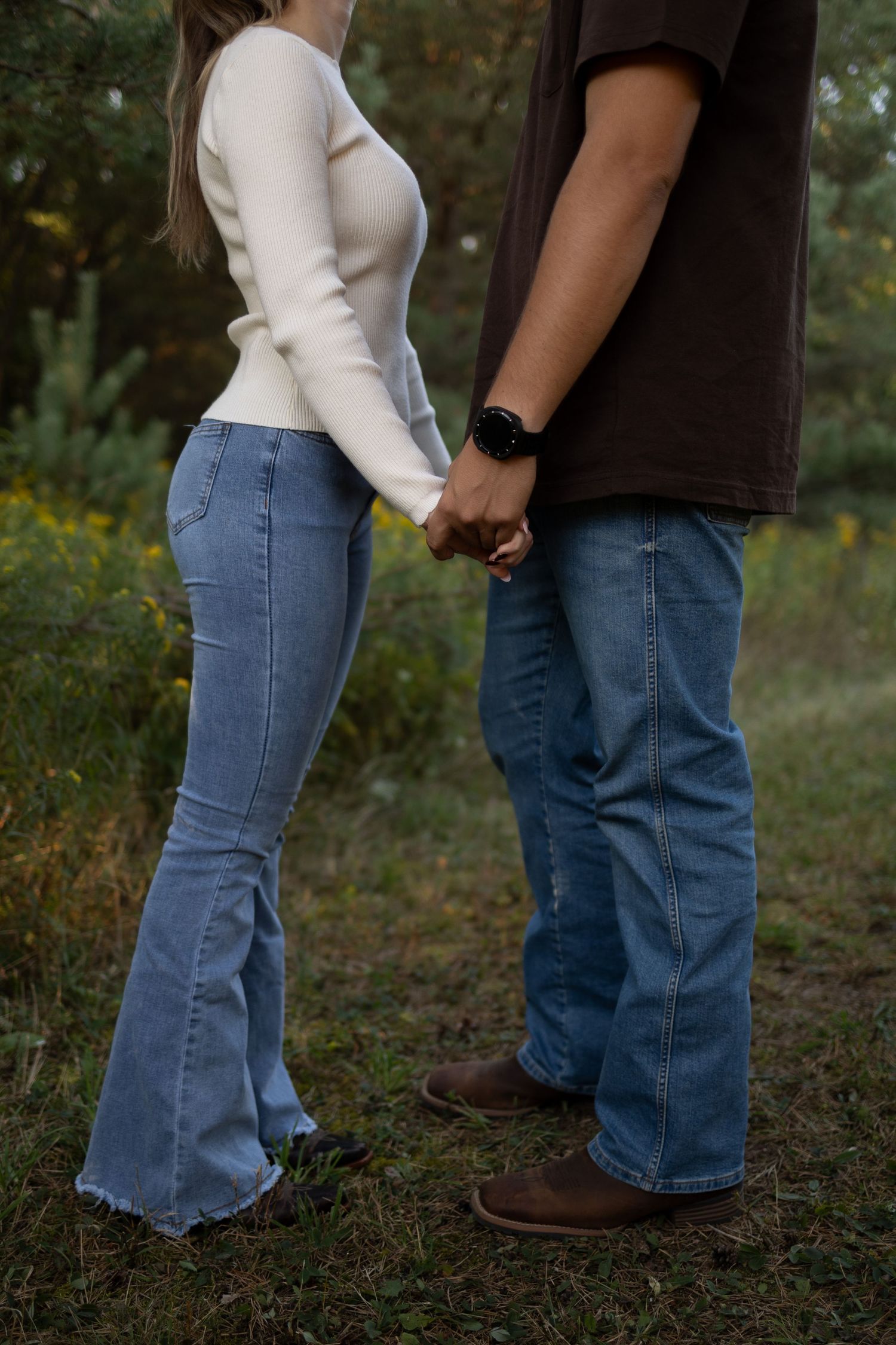Couple holding hands outdoors in jeans, woman in white top, man in brown shirt, standing in green field.