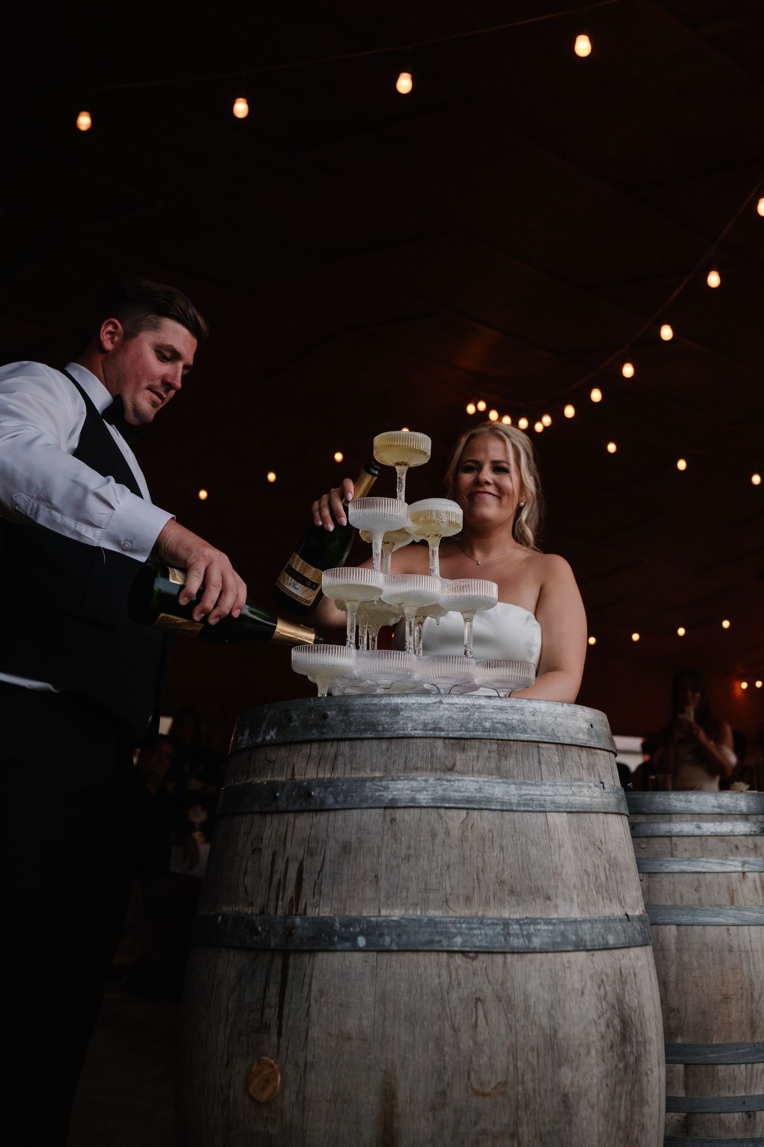 Bride and groom pouring champagne tower on wine barrel at rustic wedding venue with string lights.