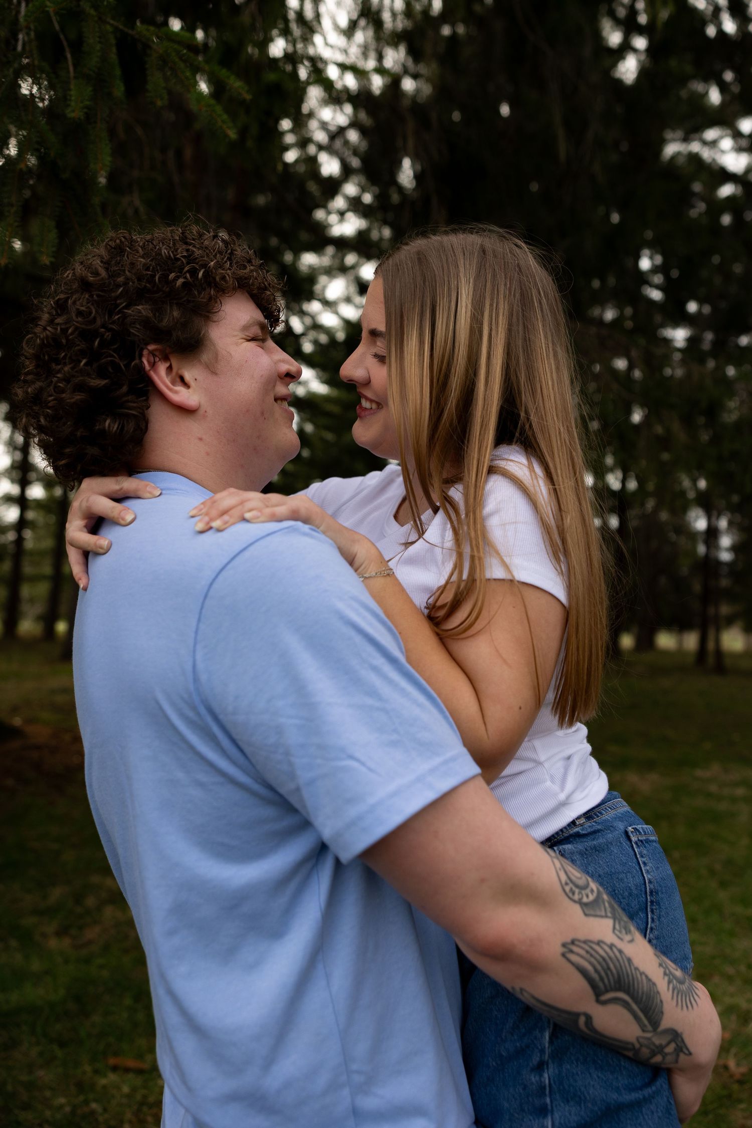 A couple embraces and smiles at each other outdoors, the man in a blue shirt and the woman in a white top and jeans.