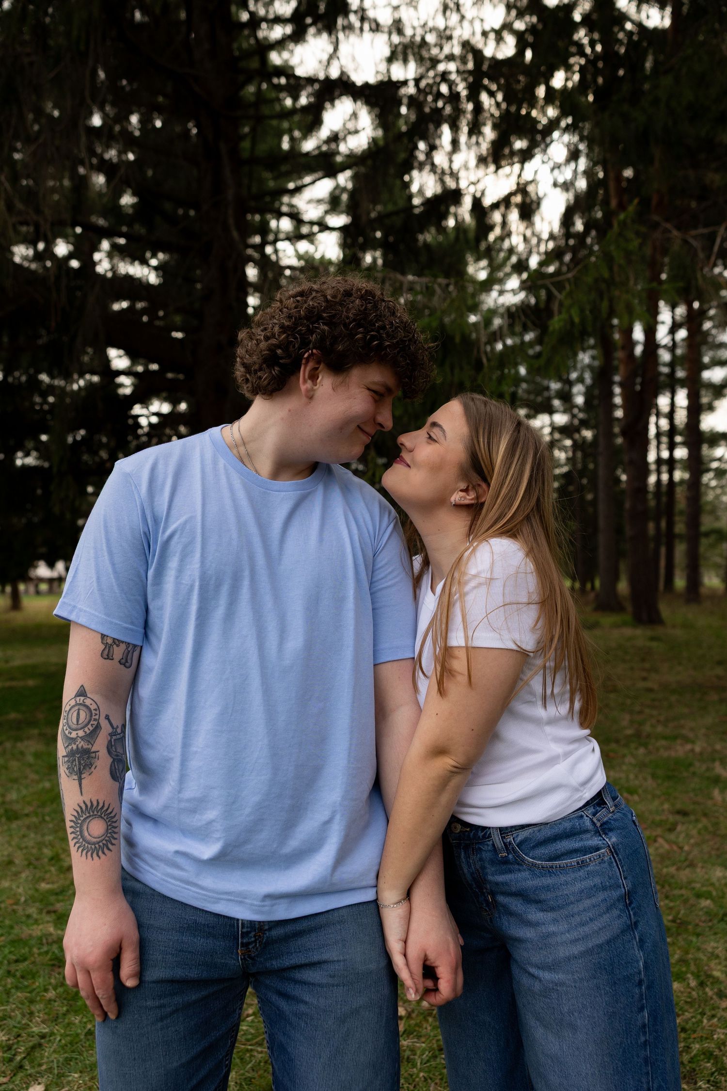 A couple sharing a romantic moment in a park, the man with curly hair and tattoos wearing a blue t-shirt and jeans.
