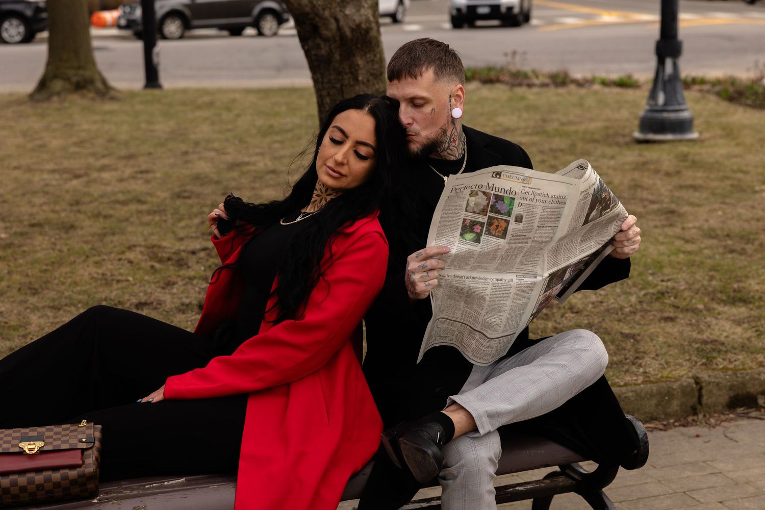 A couple sits on a park bench, woman in red coat using phone, tattooed man reading newspaper outdoors.