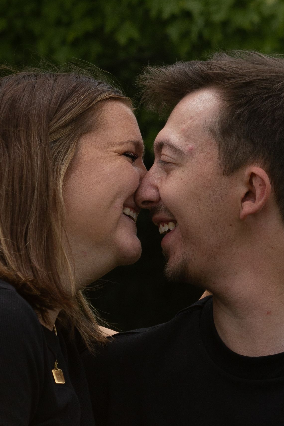 A smiling couple in black shirts share a tender nose-to-nose moment outdoors with green foliage behind them.