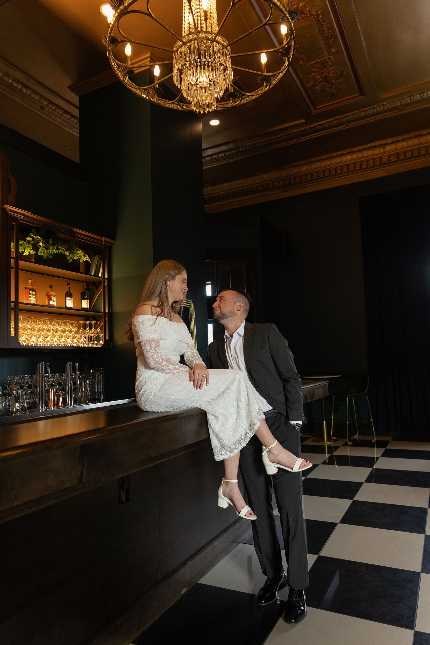 Elegant couple in a dark bar, woman in white dress seated on counter, man in suit, checkered floor, chandelier above.
