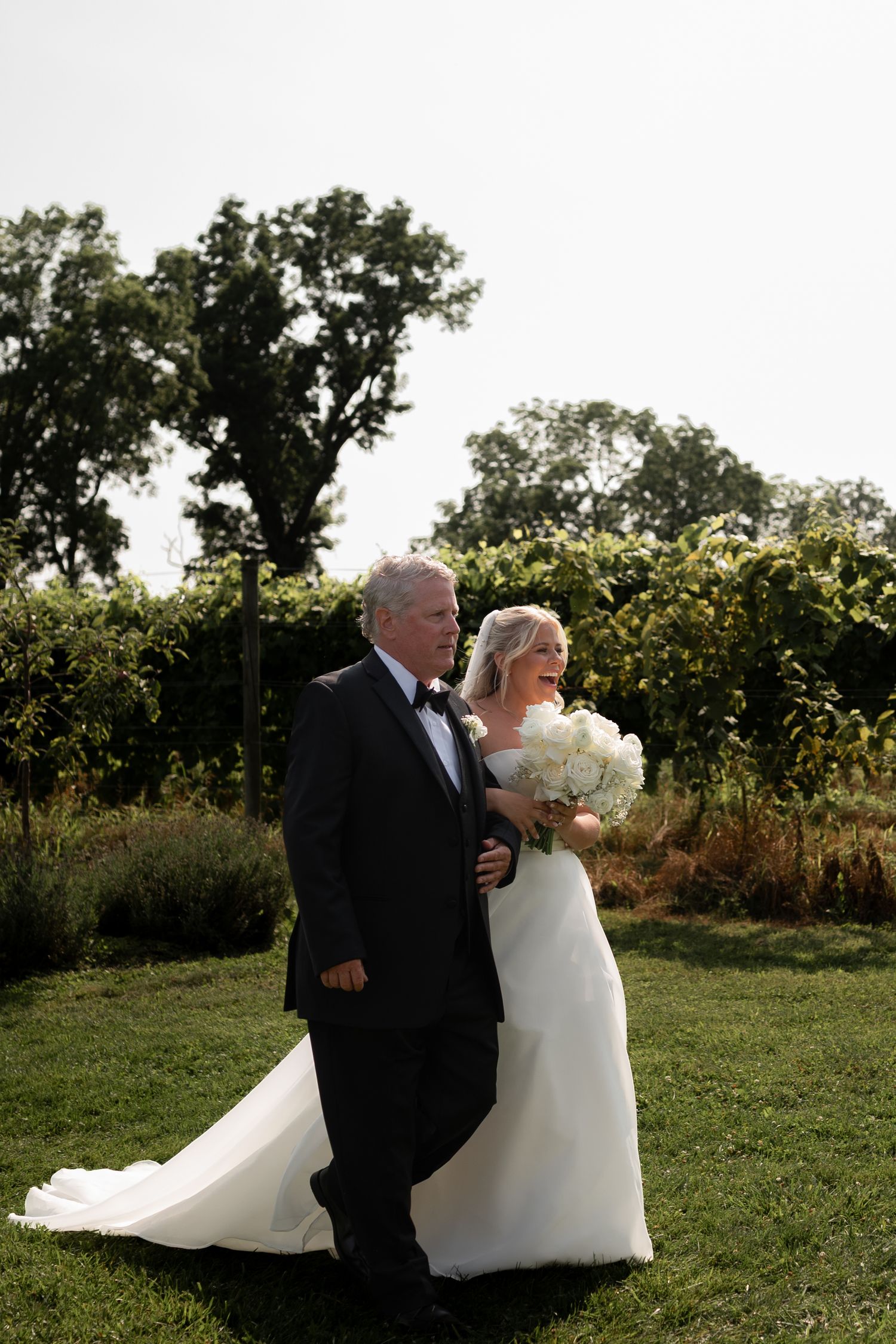 Bride in white gown holding bouquet walks with groom in tuxedo through lush garden vineyard on wedding day in Western New York
