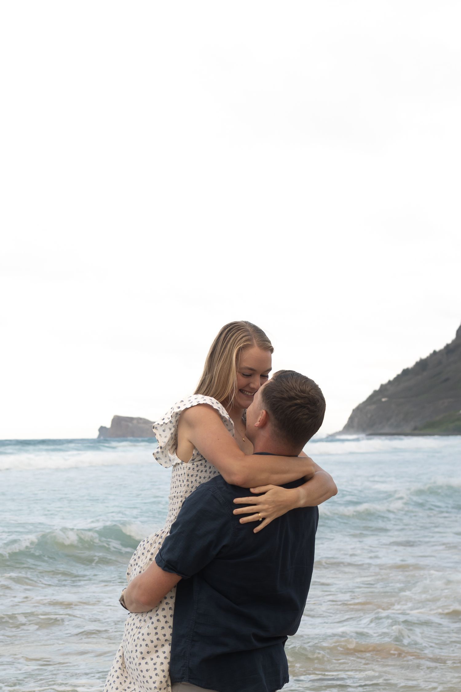 Couple embracing romantically on a beach with ocean waves and rocky cliffs in the background in Kailua, Hawaii