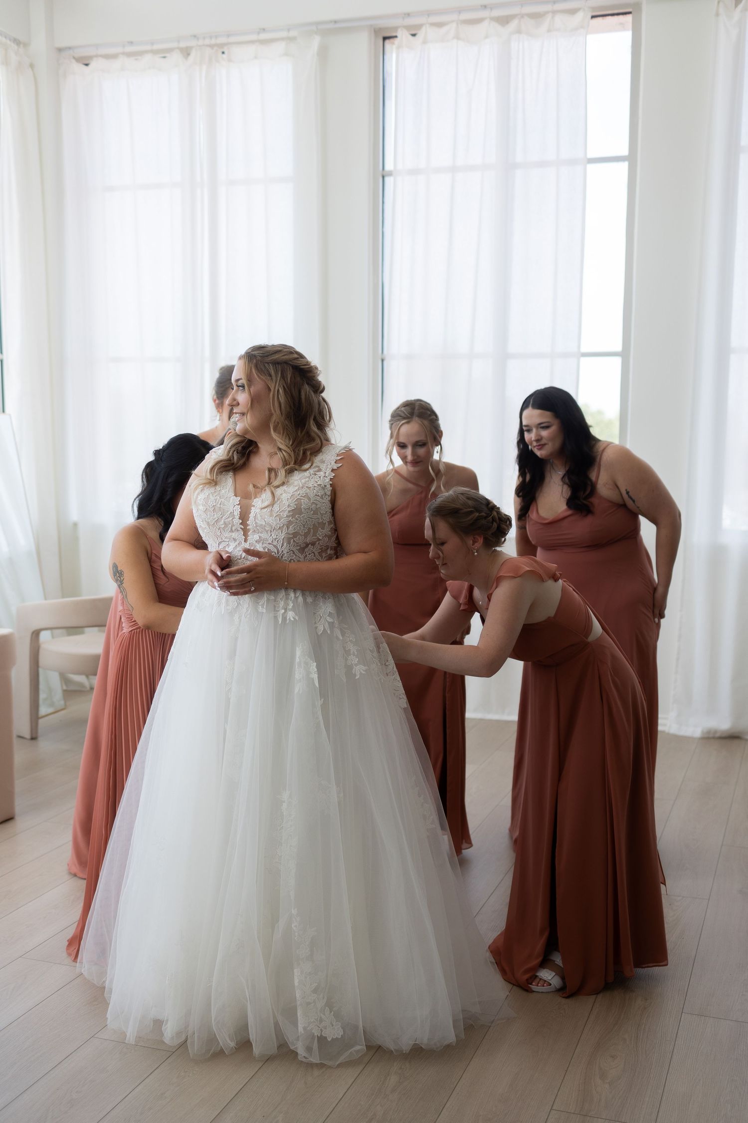 Bride in white lace ball gown surrounded by bridesmaids in rust-colored dresses being helped to get ready.