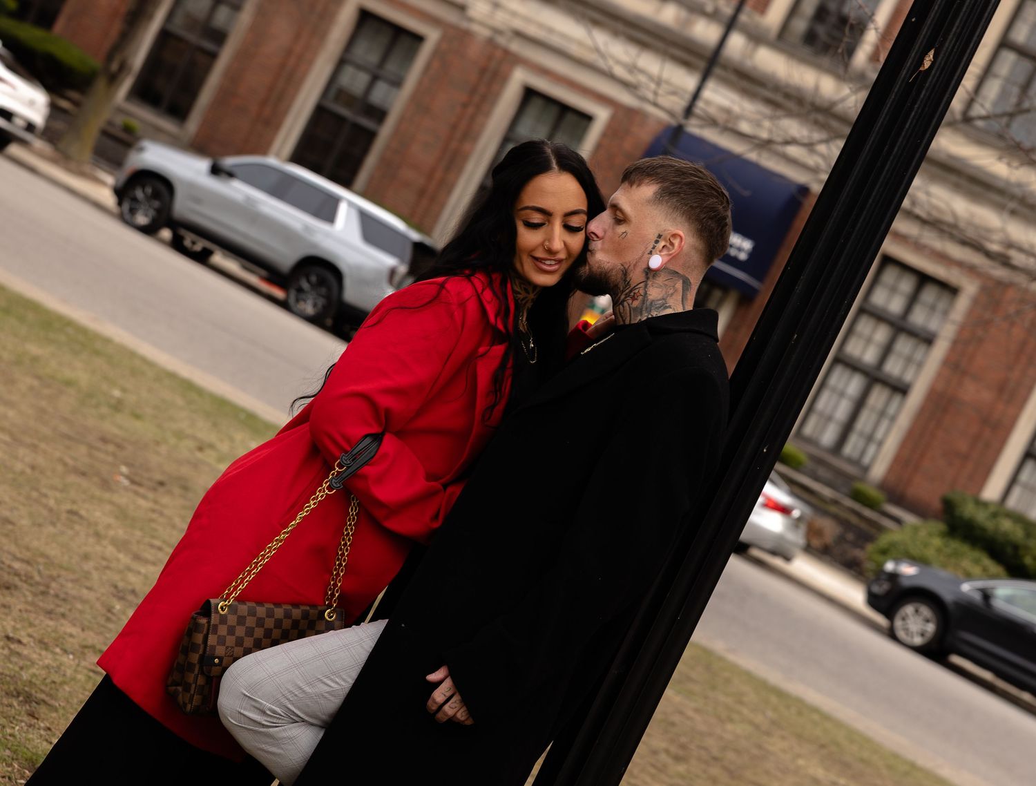 Couple sharing a romantic moment by a black pole, woman in red coat holding Louis Vuitton bag outdoors.