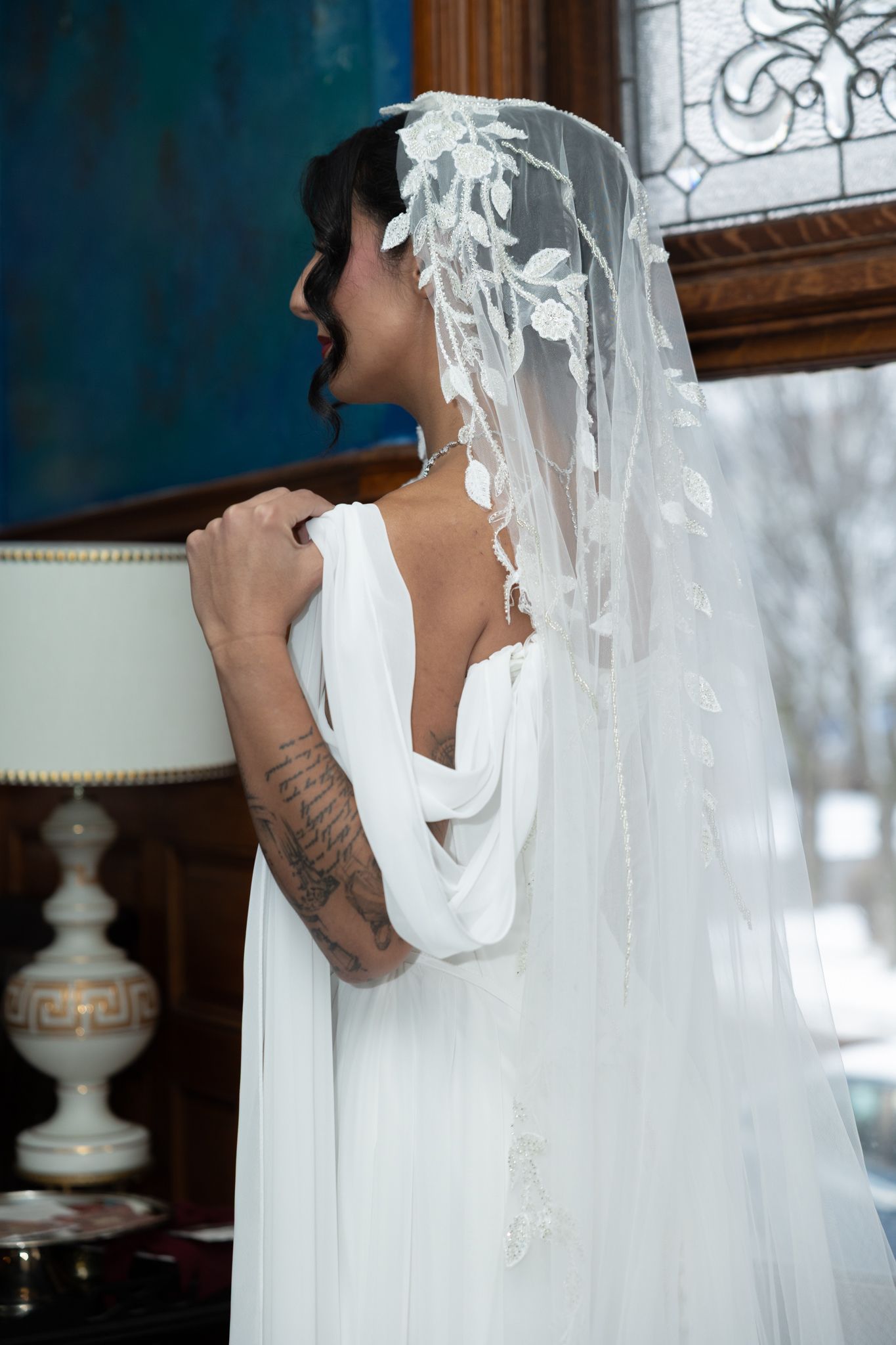 Tattooed bride in white backless gown and lace veil stands by window in elegant room.