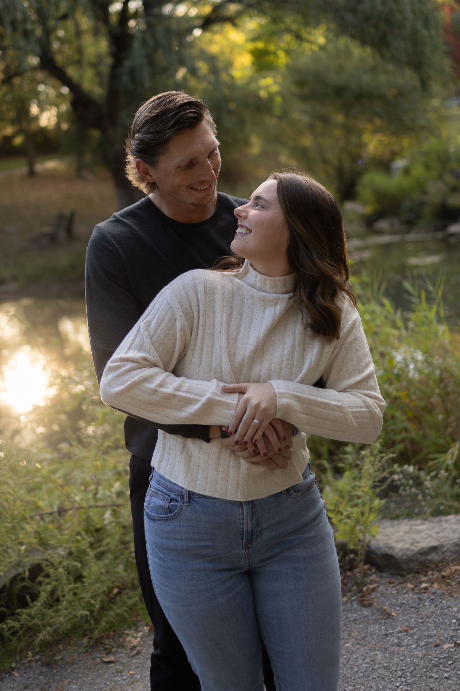 A smiling couple embraces outdoors in a park, with golden sunlight and lush greenery in the background.