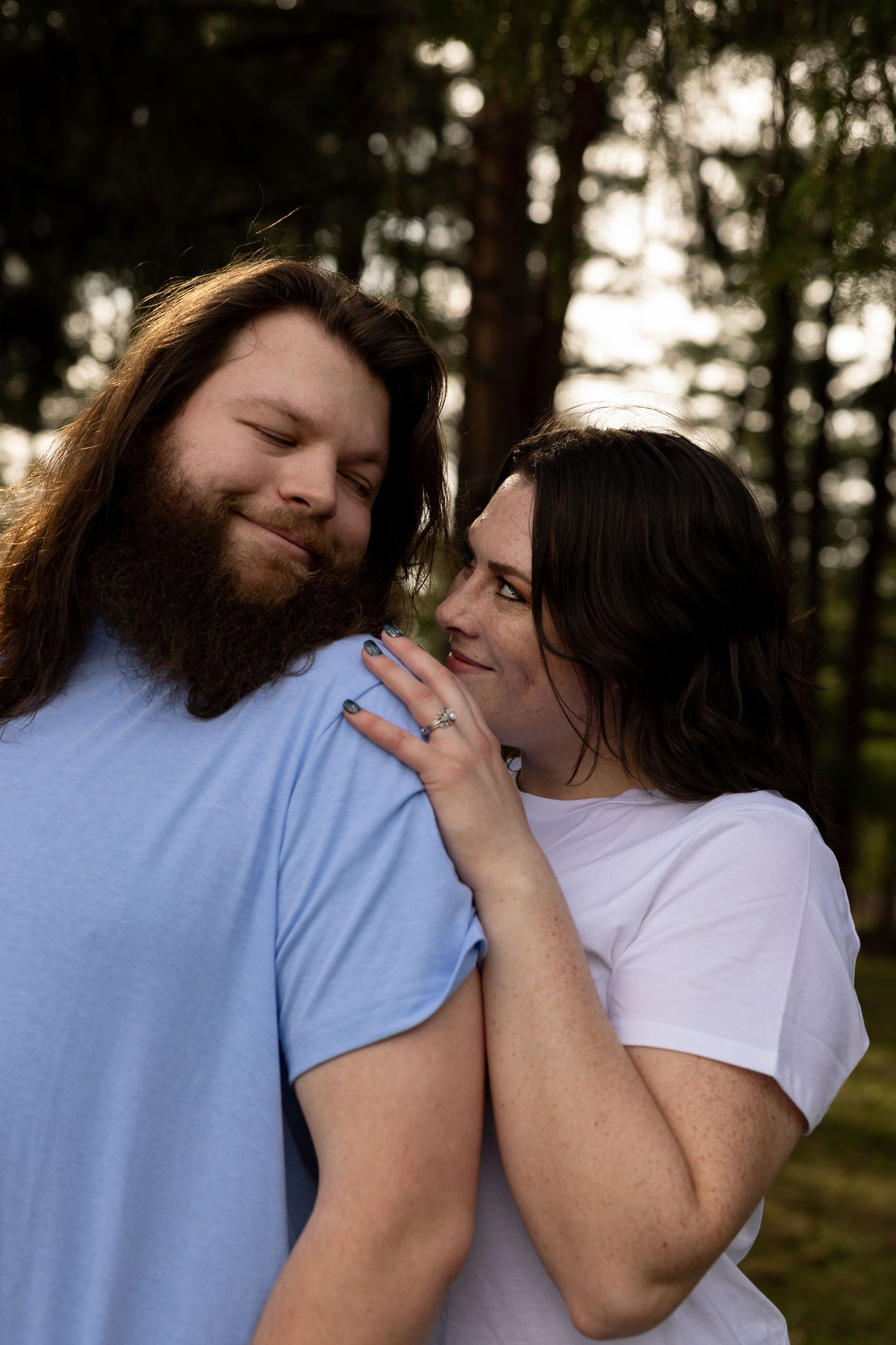 A couple shares a tender moment outdoors, woman resting her head on bearded mans shoulder among tall trees.