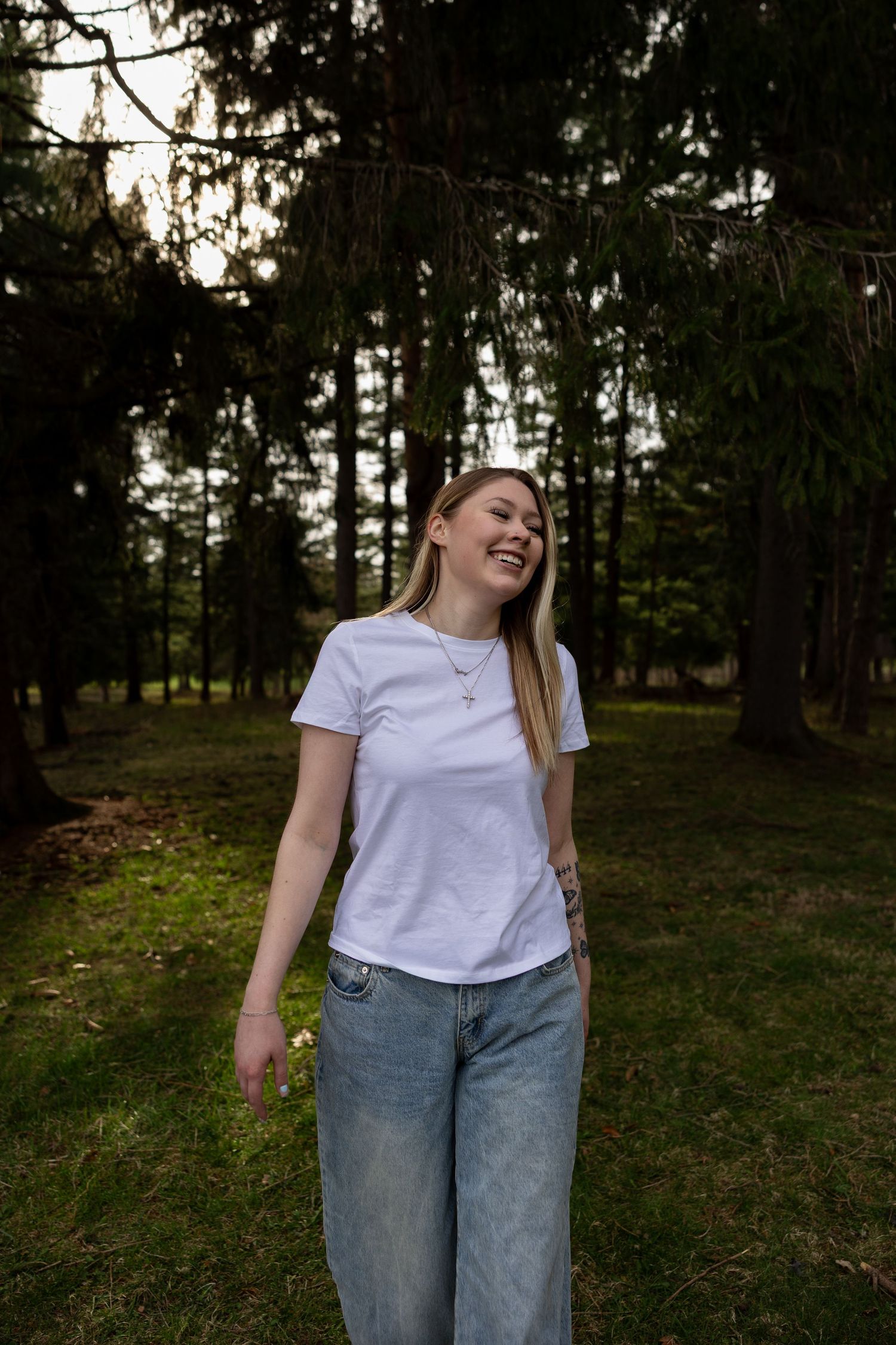 Young woman in white t-shirt and jeans smiling outdoors in a lush green forest park setting.