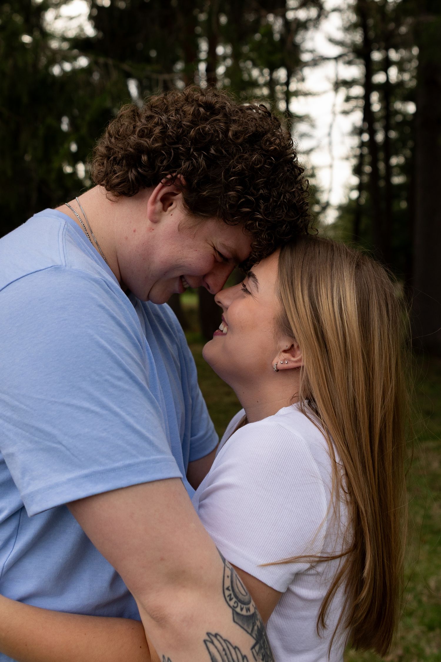 A couple smiles and touches foreheads affectionately outdoors among tall trees, wearing casual blue and white t-shirts.