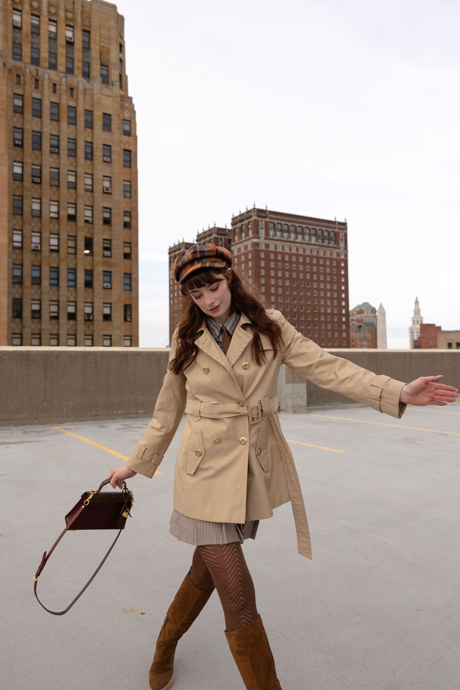 Woman in beige trench coat, plaid cap, and brown knee-high boots poses on rooftop with city buildings backdrop.