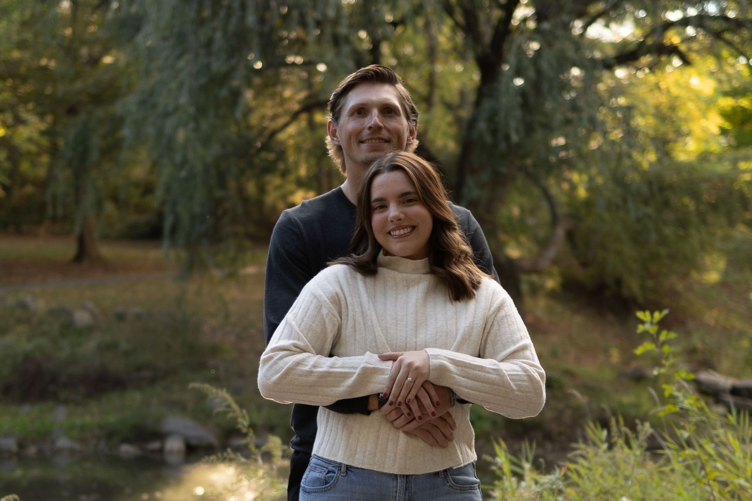 Couple embracing outdoors in a lush green park, smiling warmly near a pond with autumn trees.