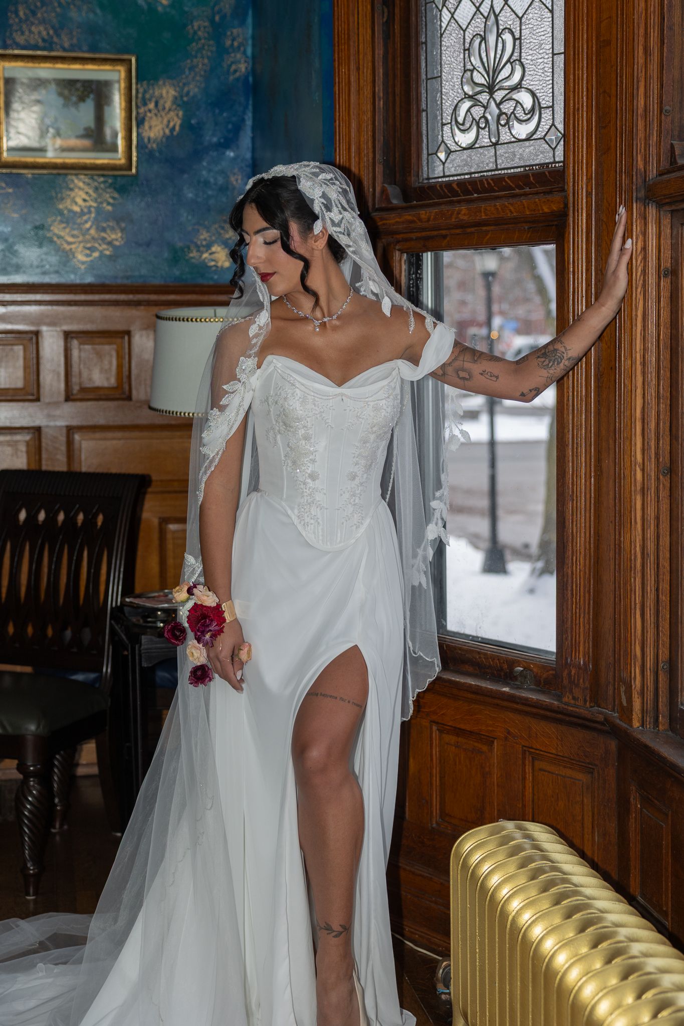 Bride in white lace corset gown with veil and bouquet poses by ornate stained glass window in elegant wood-paneled room.