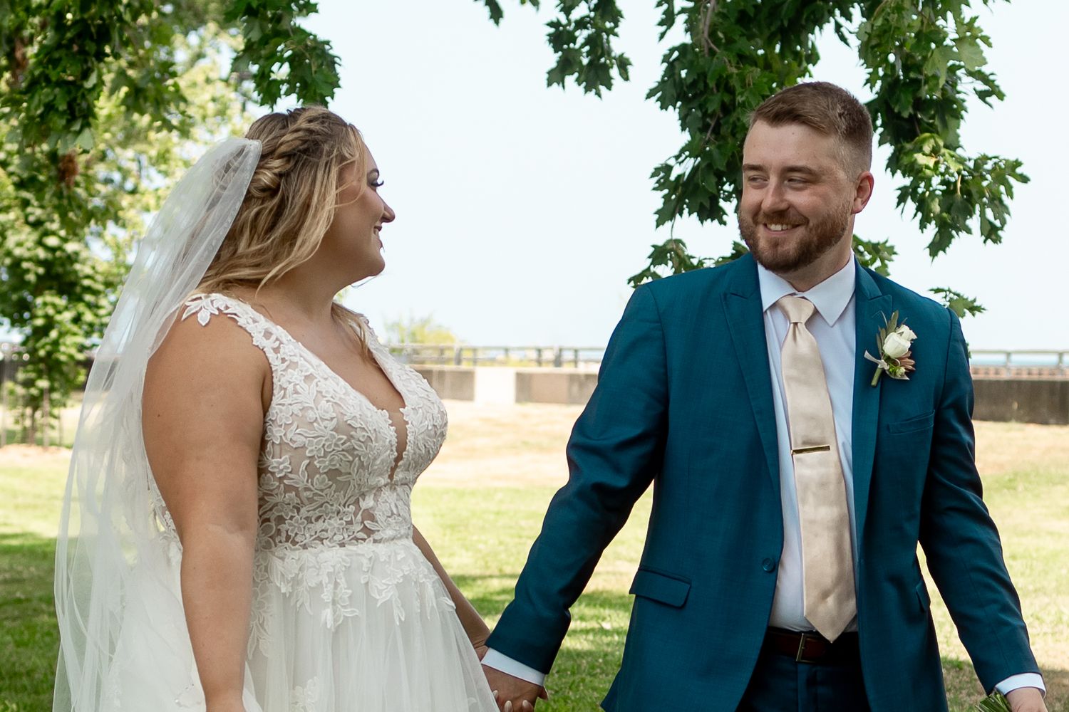 Bride in lace gown and veil holds hands with groom in teal suit outdoors on sunny wedding day.