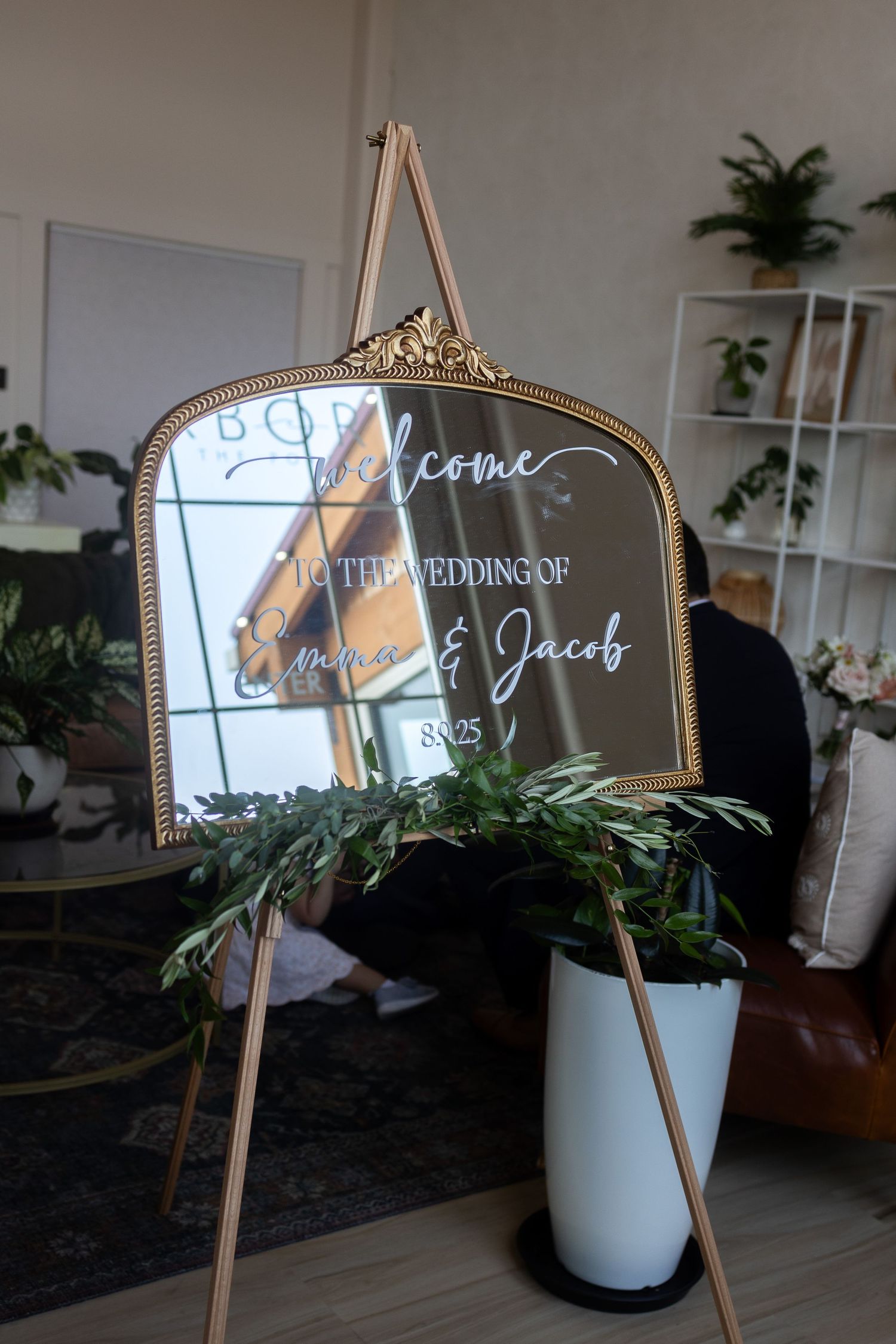 Gold ornate mirror wedding welcome sign for Emma & Jacob on wooden easel with greenery garland.