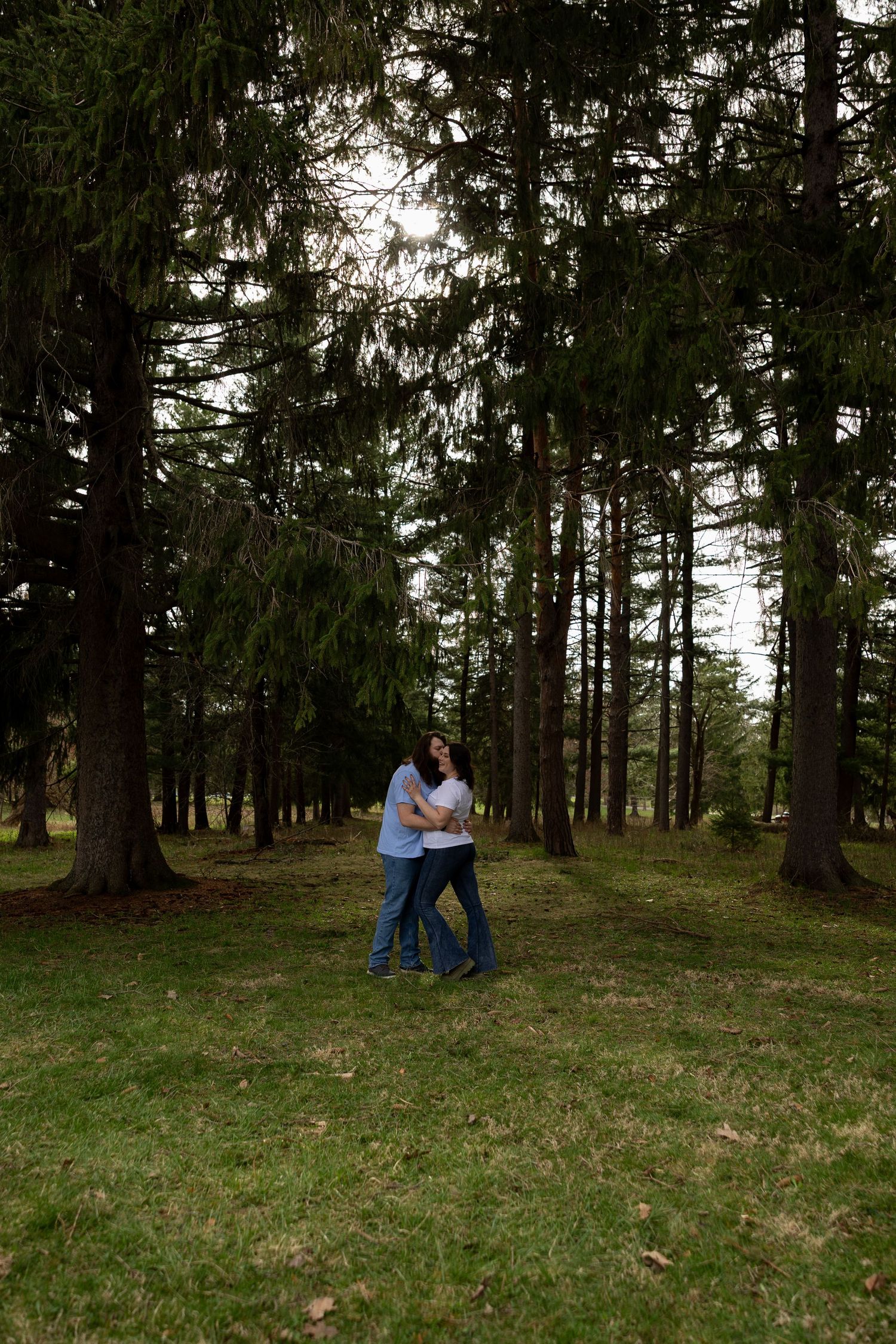 Couple embracing romantically among tall pine trees in a lush green forest park setting.