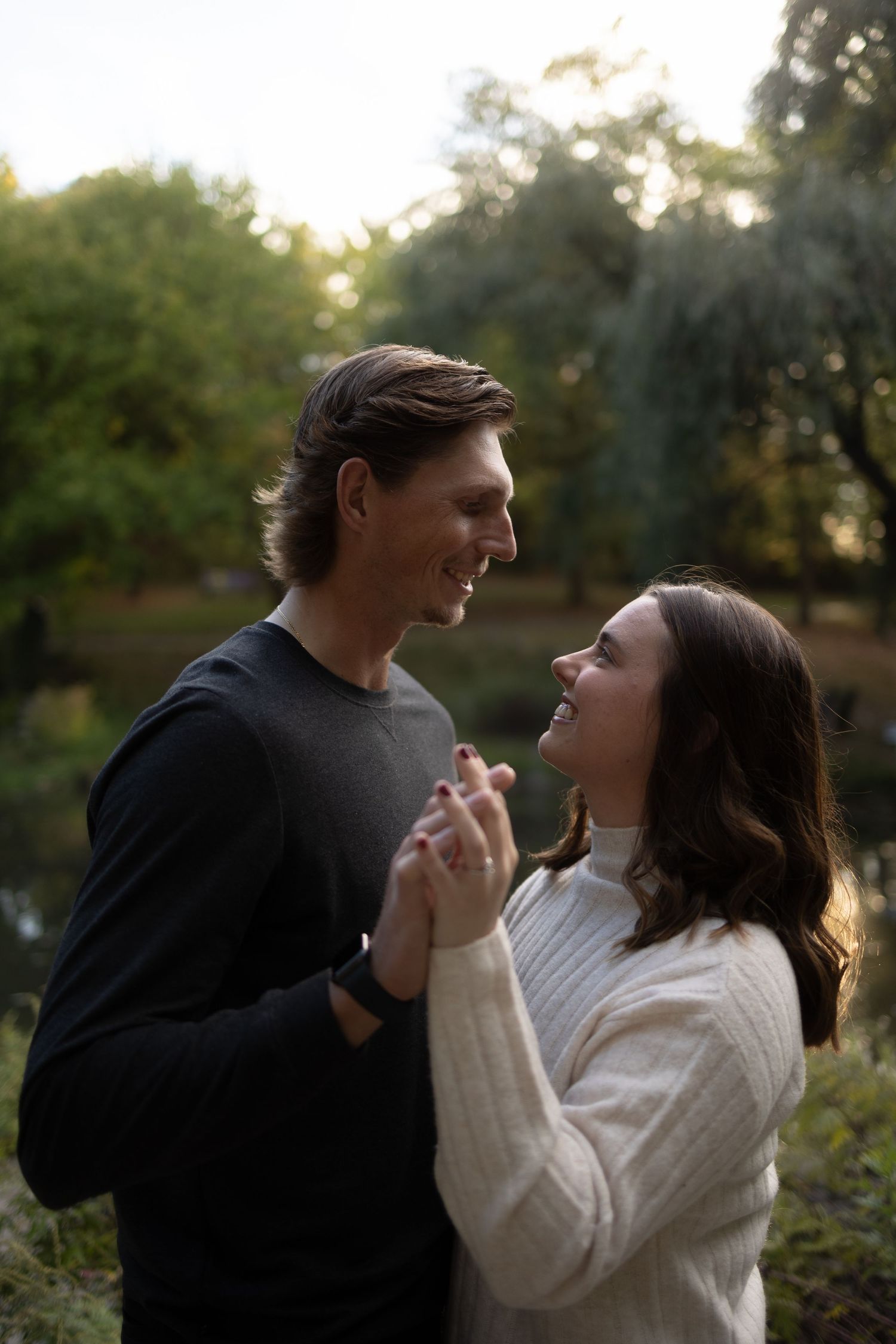 A smiling couple holds hands and gazes at each other in a lush green park at golden hour.