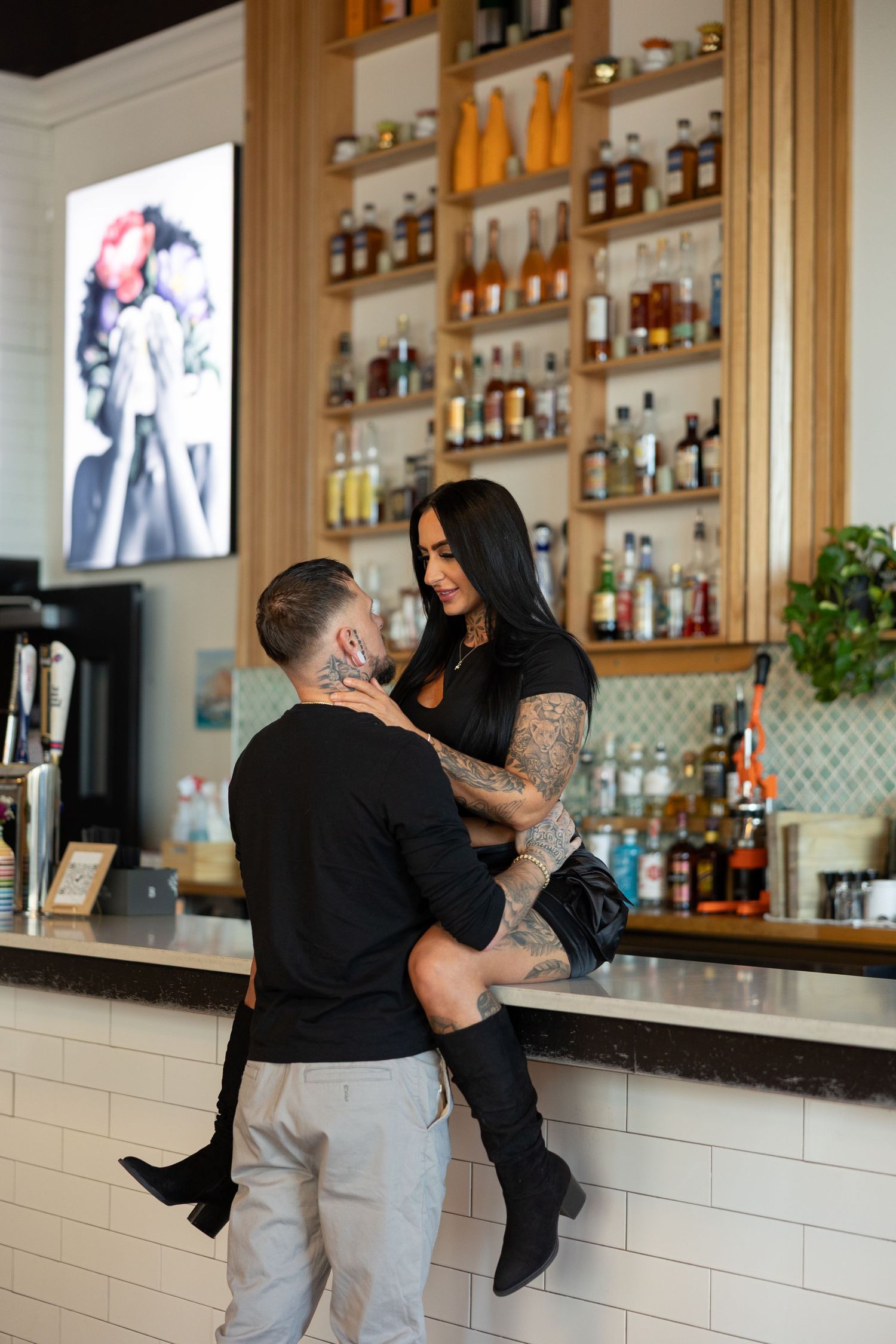 Tattooed couple sharing a romantic moment at a stylish bar, woman sitting on counter while man stands close.