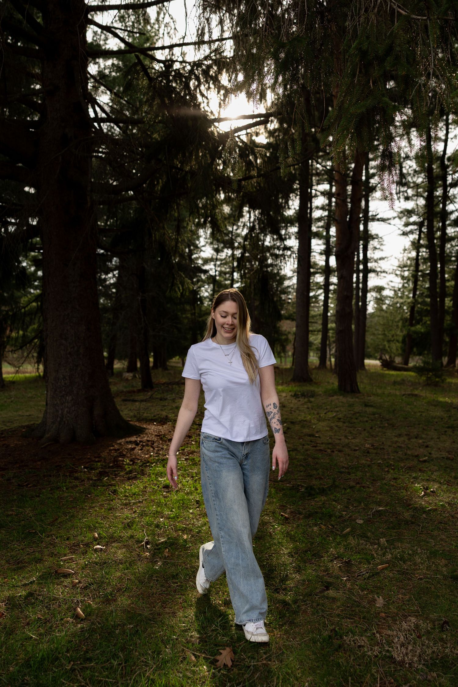 Woman in white t-shirt and jeans walking through a sunlit pine forest on green grass.