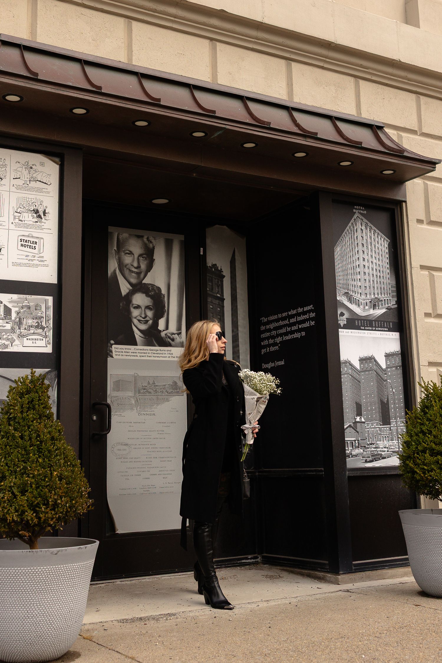 Woman in black coat holding white flowers stands outside a historic building with vintage celebrity photos on display.