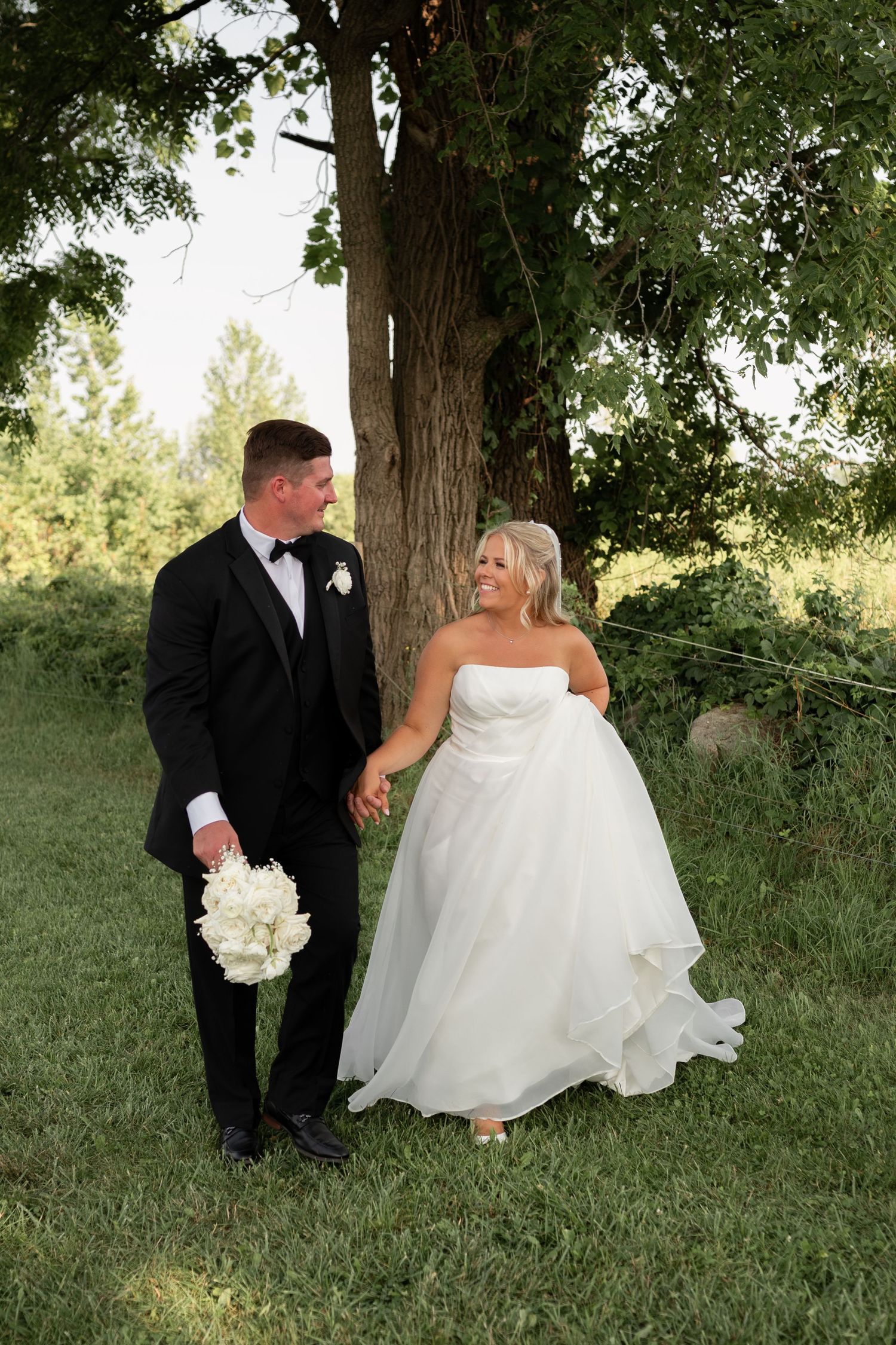 A bride and groom holding hands and smiling at each other outdoors near a large tree on their wedding day.