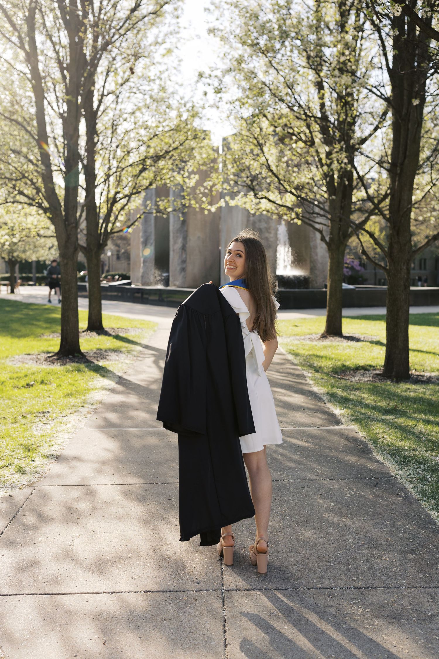 Graduate walks along tree-lined path carrying graduation gown.