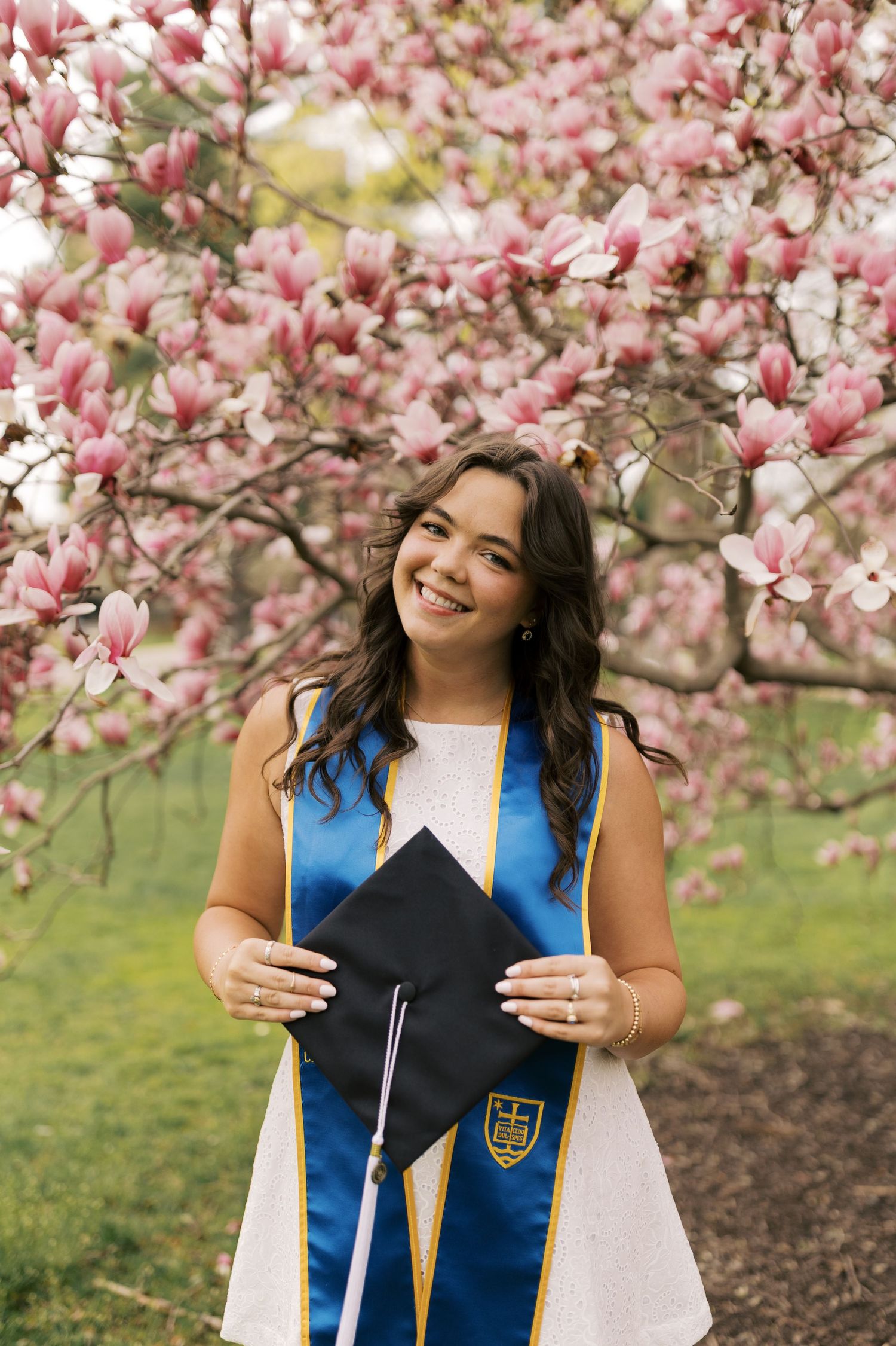 A graduate poses with their cap under blooming pink magnolia trees during spring graduation season.