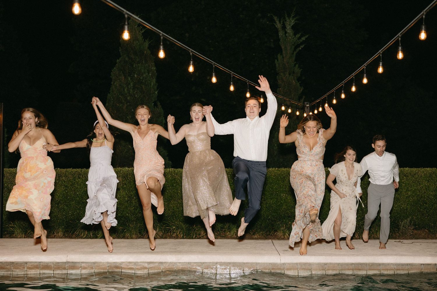 Group of wedding guests dancing and jumping under string lights at night.